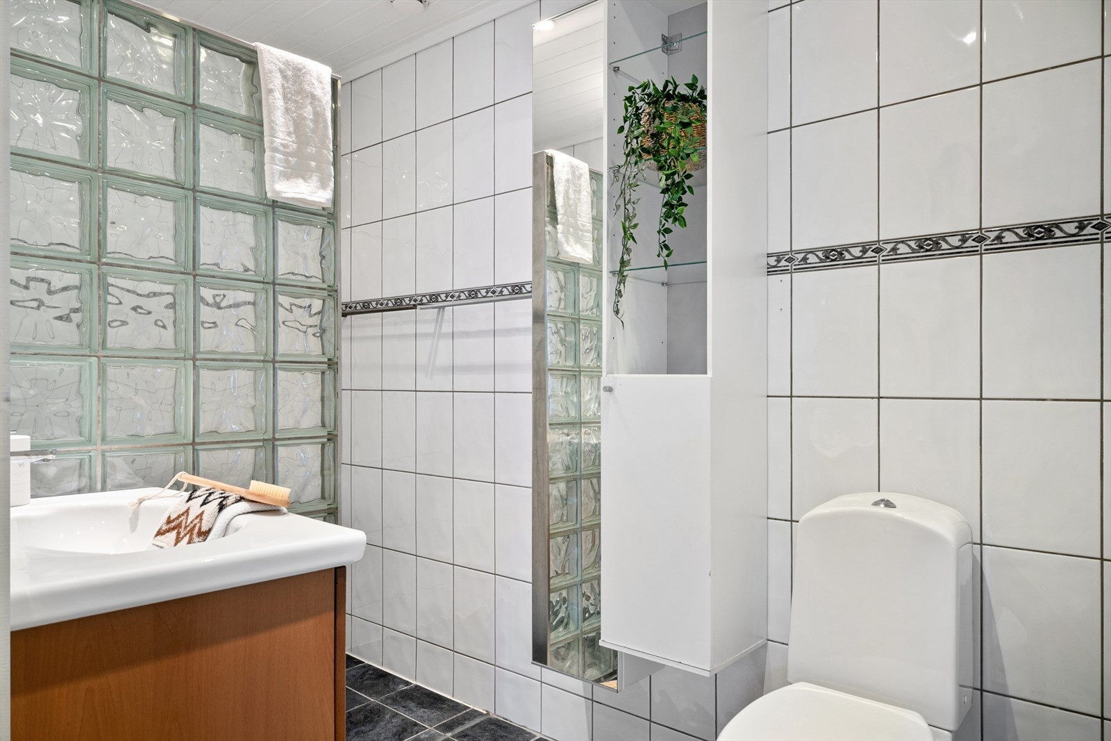 Badet har fliser på gulv og vegger. Badet er innredet med servant i baderomsmøbel, toalett og dusjnisje med glassbyggerstein. Galleribilde