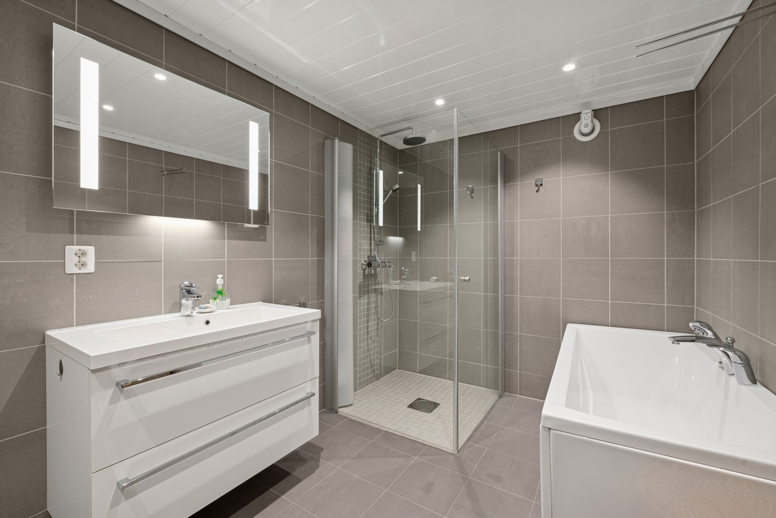 Rommet er pent gjennomført med flislagte vegger og gulv. Det er behagelig varme i gulvet og spotbelysning i taket. Rommet er av god størrelse, og inneholder både dusjnisje og badekar til glede for store og små. Galleribilde