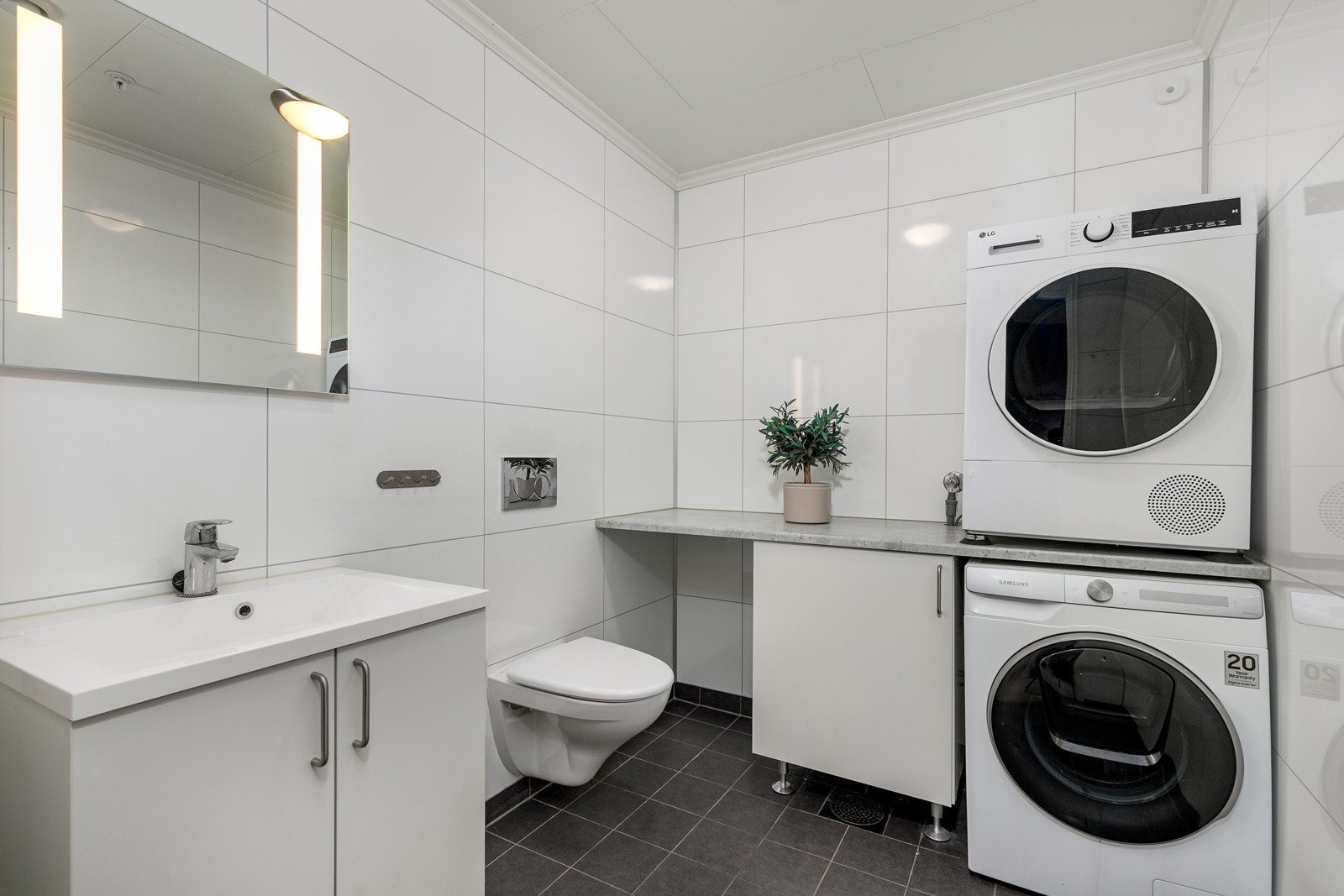 Lyst og praktisk vaskerom. Vaskerommet har opplegg for vaskemaskin og tørketrommel i søyle, samt benk med skap. Fliser på gulv og baderomsplater på vegger. Galleribilde