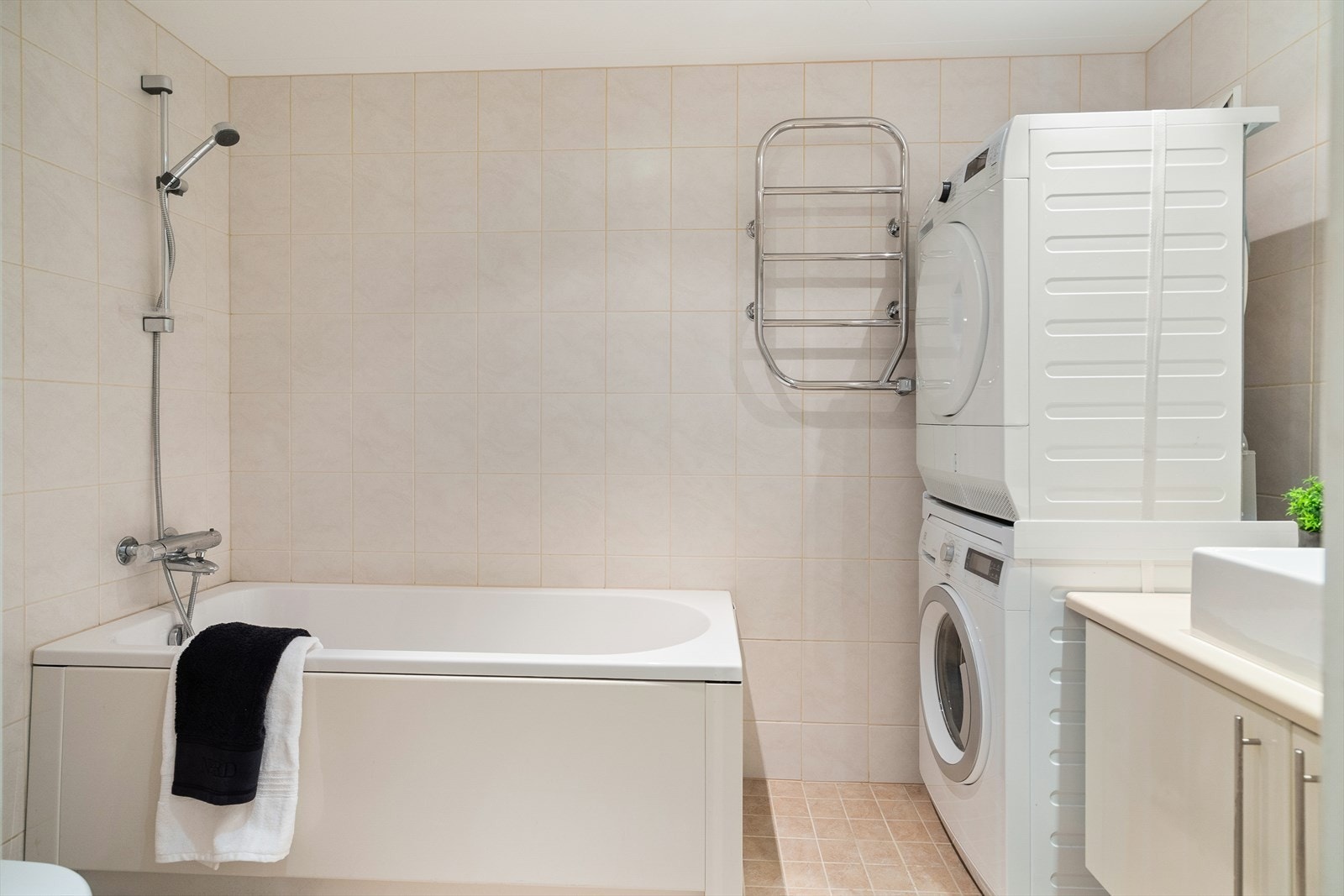 Delikat bad med badekar og moderne innredning bestående av badekar med dusjløsning, servant med underskap, speil med belysning, veggmontert toalett, håndkletørker og opplegg for vaskemaskin. Galleribilde