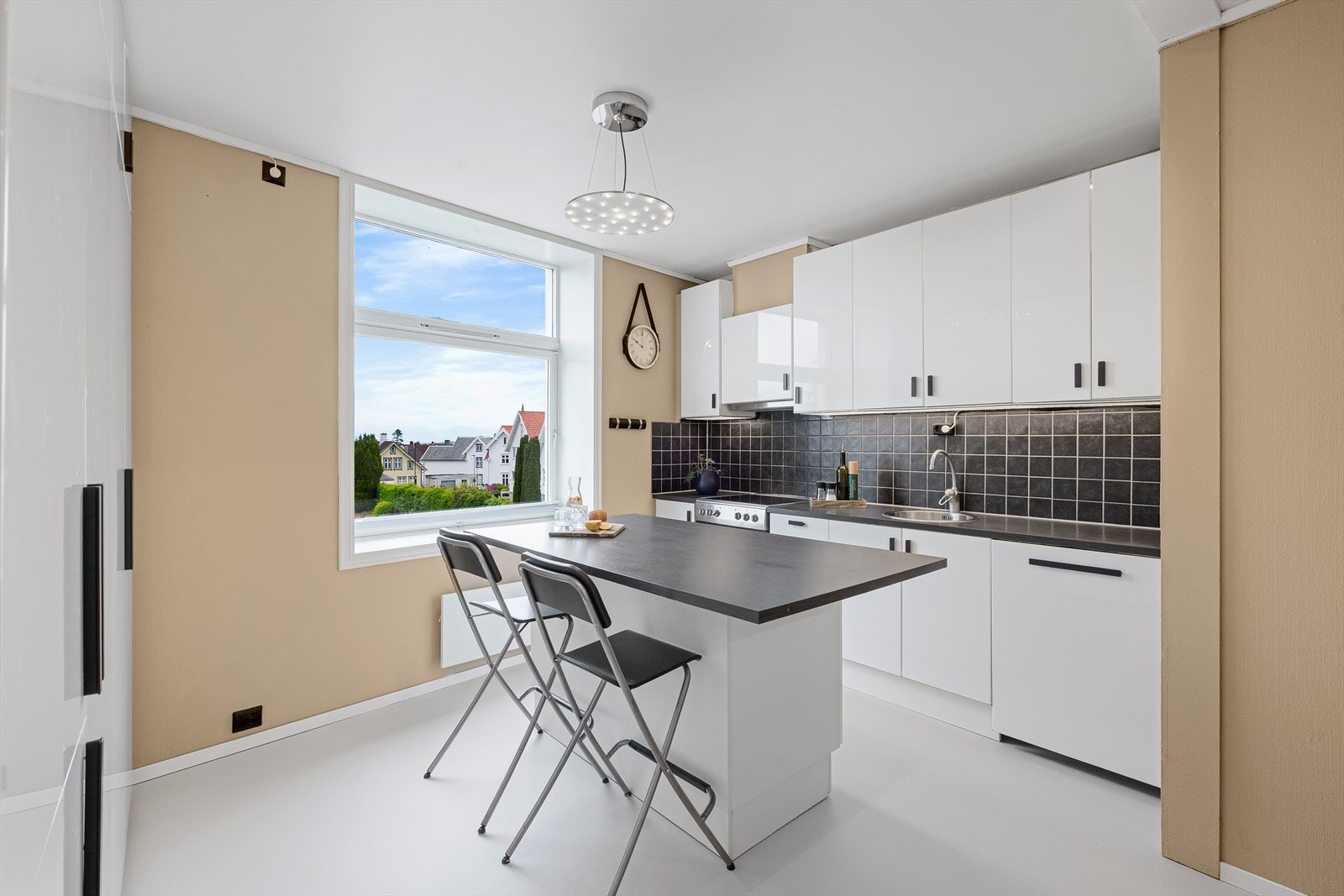 Kjøkkenet har en tilbaketrukket plassering fra den sosiale sonen i et eget rom, og består av omfangsrik kjøkkeninnredning med hvit høyglans fronter og mørk laminat benkeplate. Galleribilde