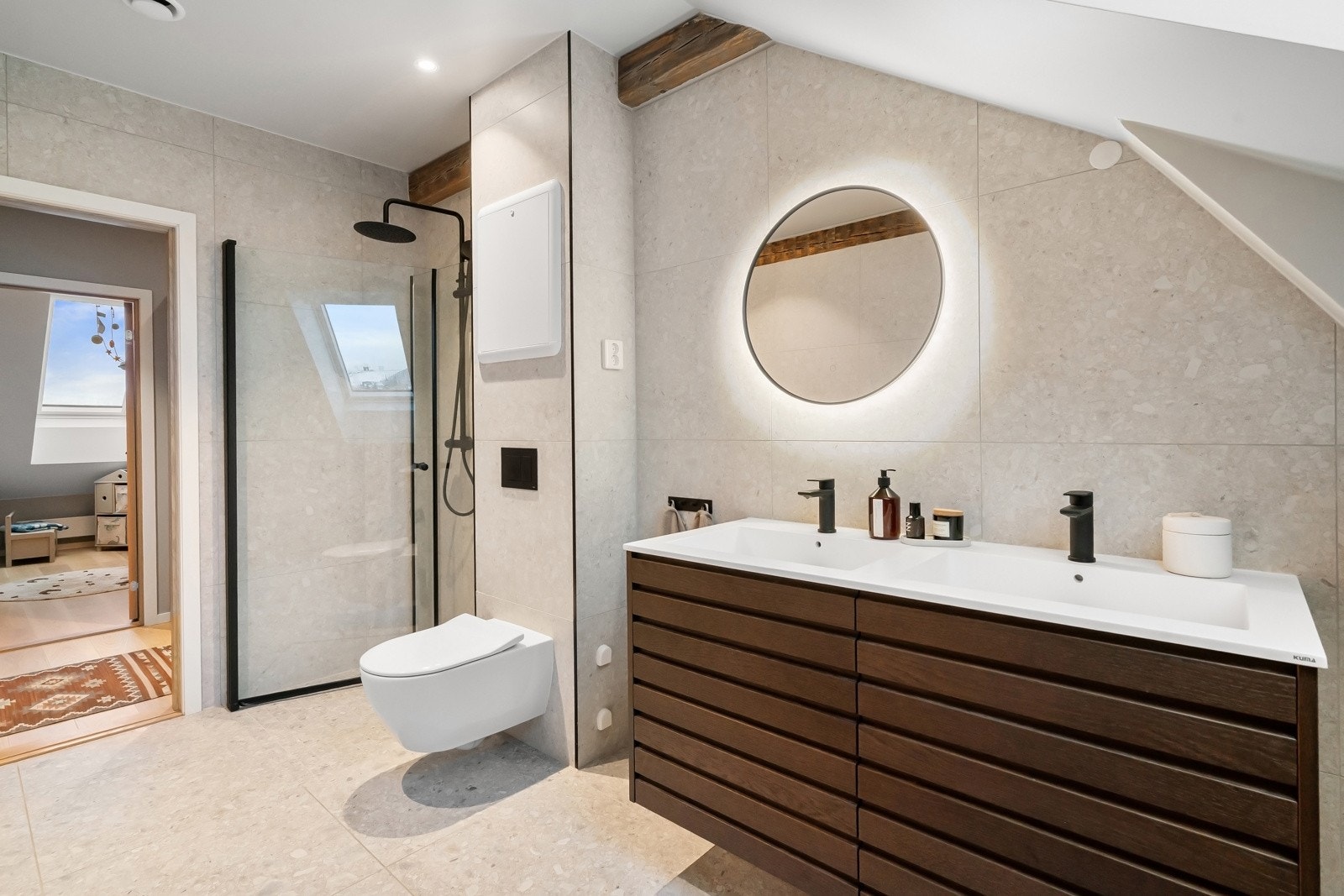 Baderomsinnredningen har mørke, profilerte eikefronter og består av en toppmontert dobbelservant på skuffeseksjon og et stort, rundt speil. Videre har badet vegghengt toalett fra Villeroy & Boch med DirectFlush, innbygningssisterne fra TECE. Galleribilde