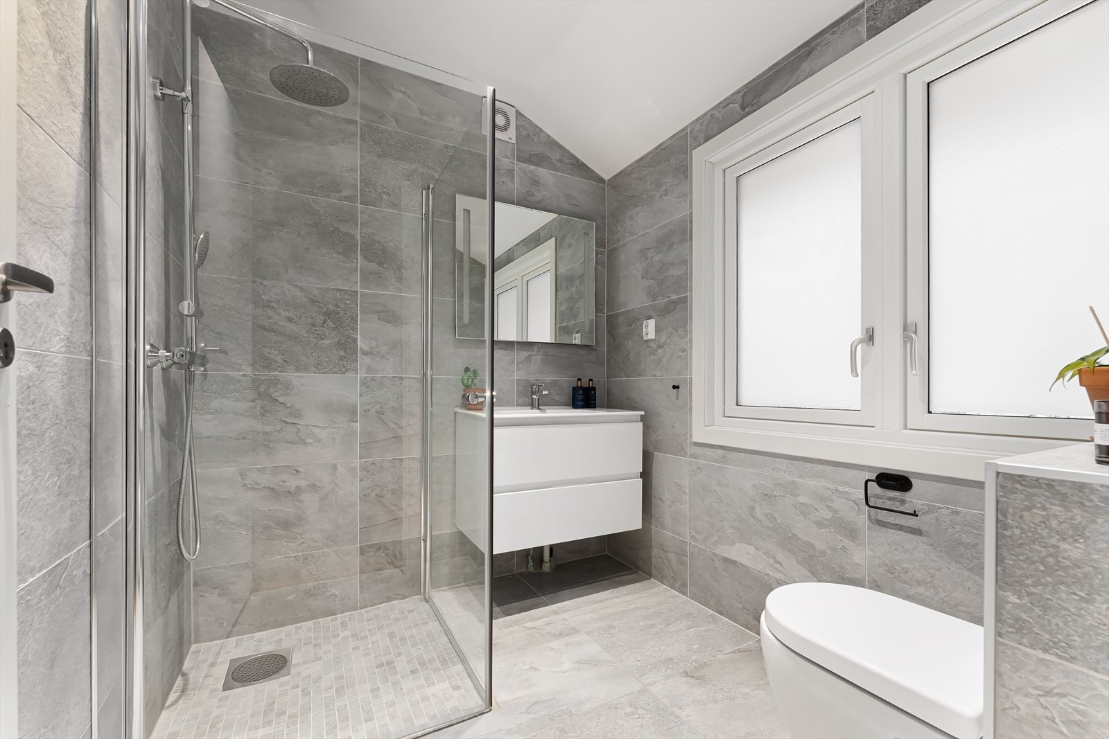 Bad i 2. etasje med regnfalldusj, innsvingbare dører i glass, veggmontert toalett, servant med skuffer og ledspeil. Galleribilde