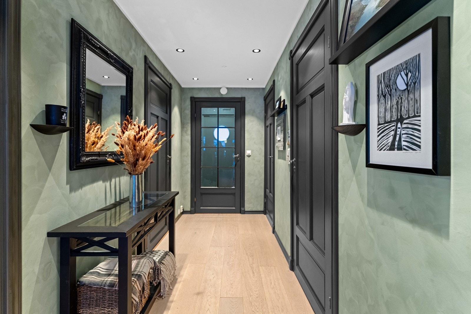 Gangen ble oppgradert i 2022, med ny kalkmaling, ny 1-stavs parkett og nye downlights i en senket himling. Veggene er malt i mørkgrønt, og gangen glir sømløst over i stuen - holdt i den samme lune fargen. Galleribilde