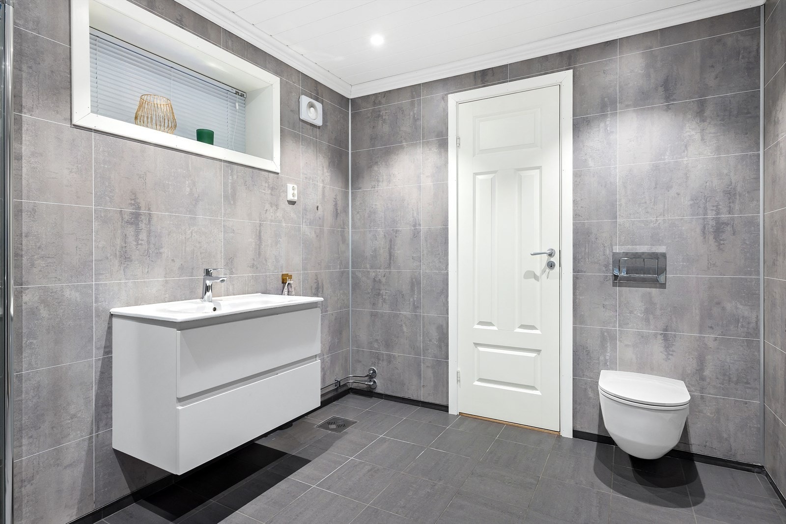 Badet inneholder dusjhjørne med svingbare glassdører og regnfallsdusj, heldekkende servant med skuffeseksjoner, vegghengt toalett og høyskap. Det er opplegg for vaskemaskin Galleribilde