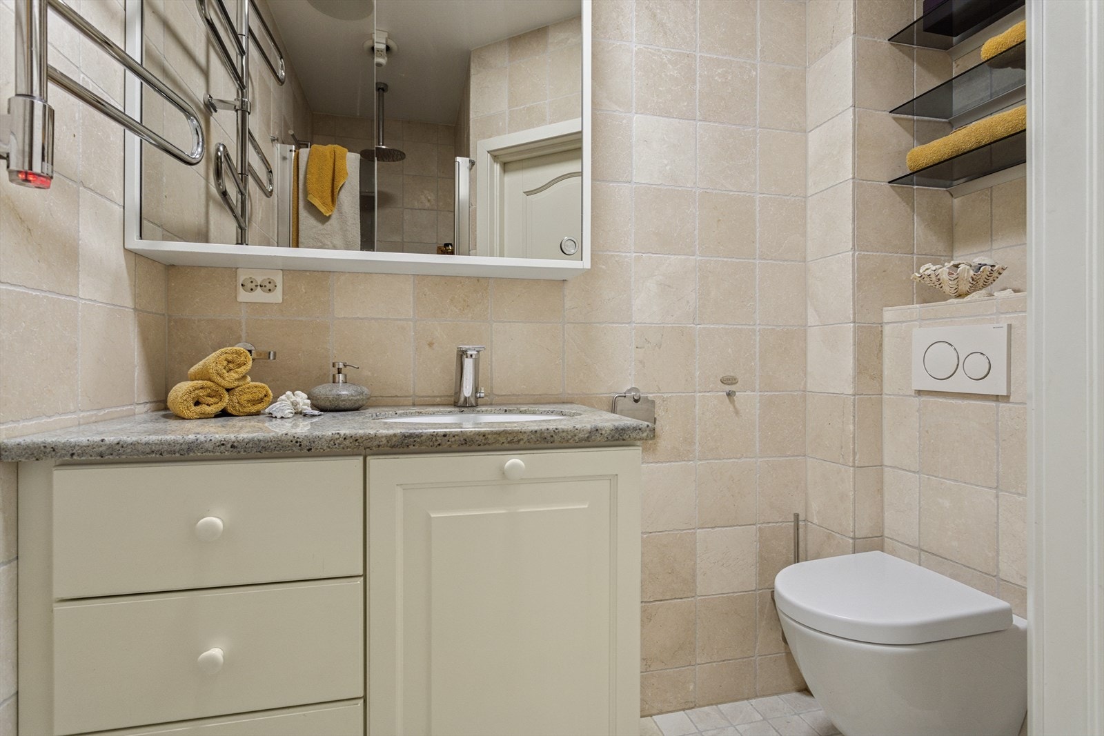 Badet har et vegghengt toalett fra Geberit med kullfilter og Bluetab, dusjdører i glass med dusjgarnityr på vegg, vaskefat fra Villeroy & Boch, håndkletørker med regulerbar varme, og en benkeplate i granitt. Galleribilde