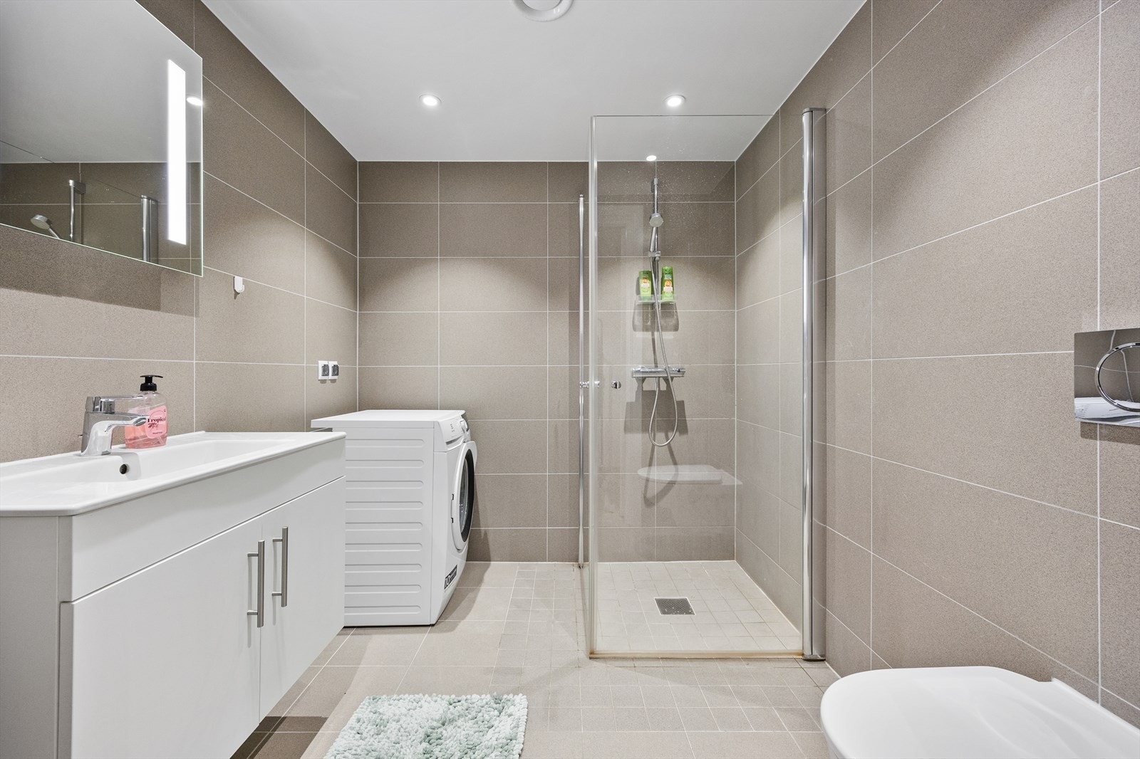 Badet har grå fliser på vegger og gulv samt gulvvarme og opplegg for vaskemaskin. Galleribilde