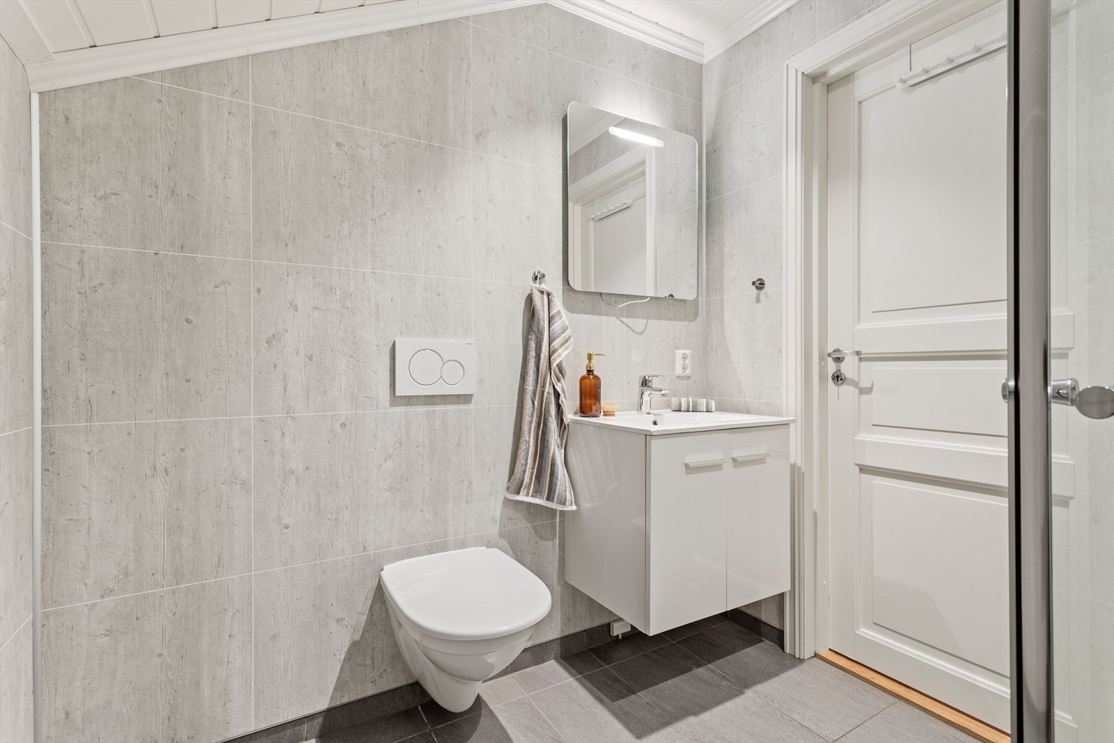 Det er nytta flis på golv og baderomspanel på veggar. I taket er det MDF-panel. Innreiing består av dusjhjørne, servant med underskåp og veggmontert klosett. Galleribilde