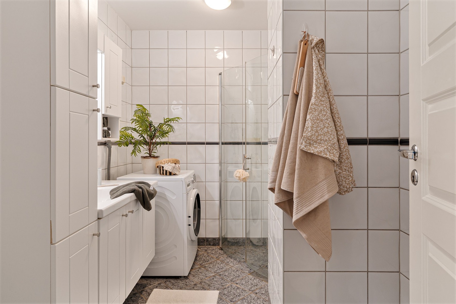 Flislagt baderom med varmekabler i gulv, dusjhjørne med innfellbare dører, opplegg for vaskemaskin, servant med skuffeseksjon og overskap, og toalett. Galleribilde