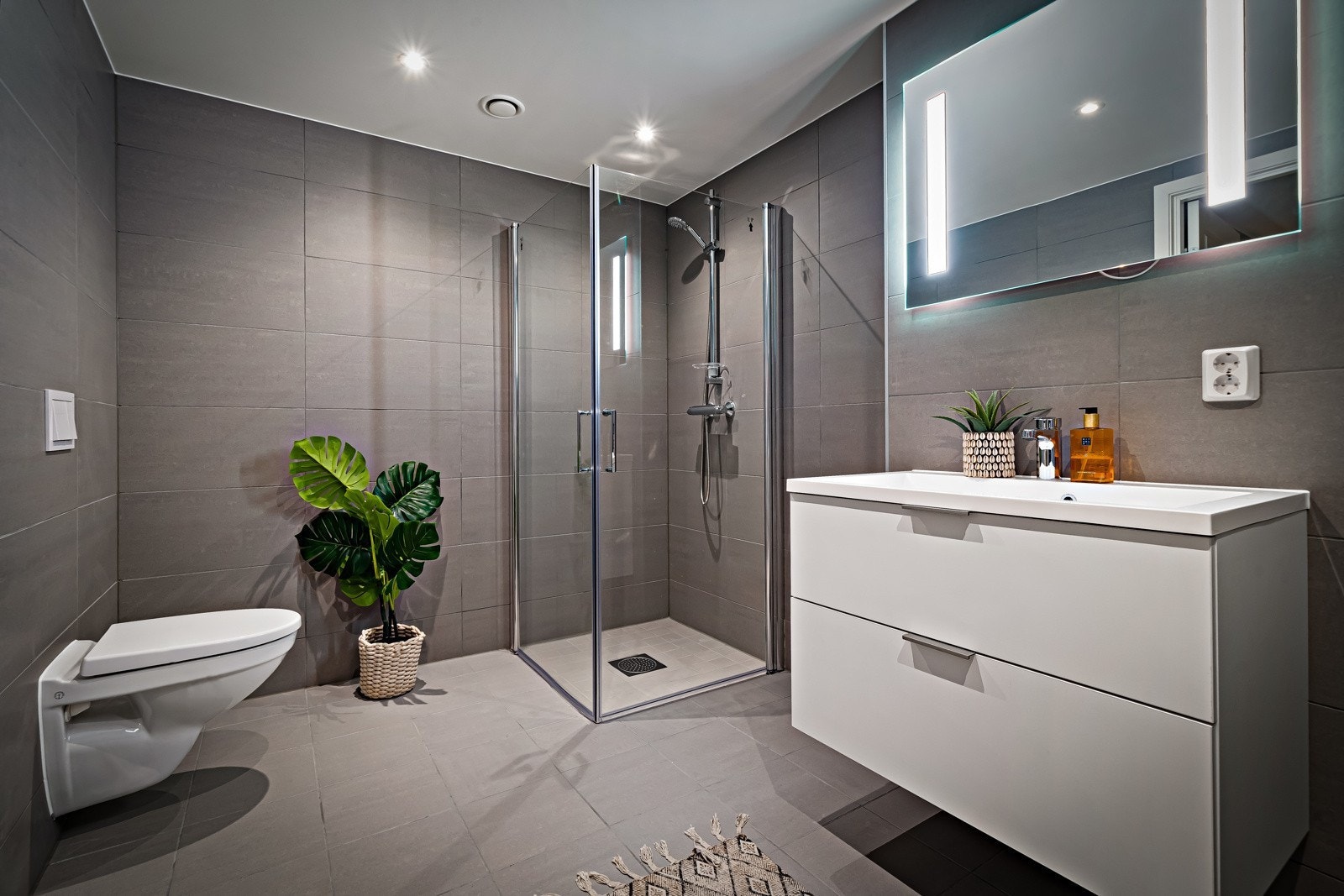 Badet er innredet med vegghengt toalett med innebygd sisterne kasse, servant på vegghengt servantskap og vegghengte dusjdører i glass Galleribilde