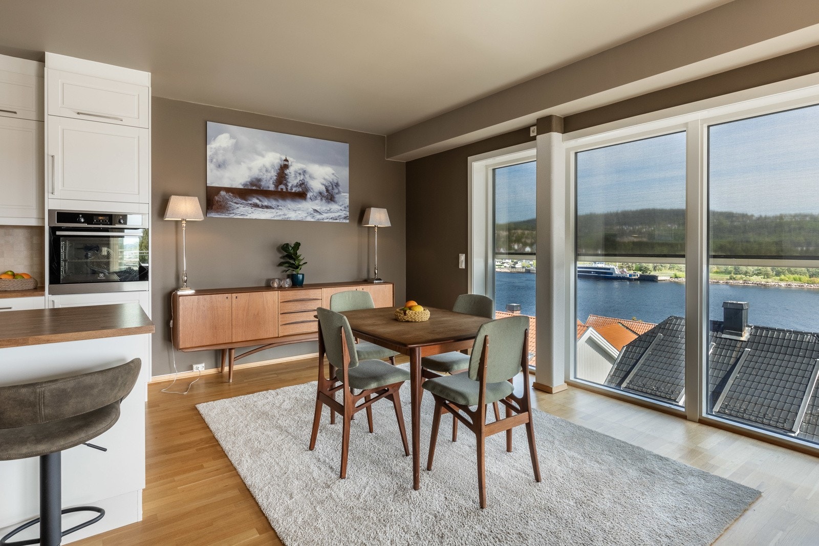 Lys stue med spisebord, store vinduer og utsikt mot vannet. Åpen kjøkkenløsning med barstoler. Galleribilde