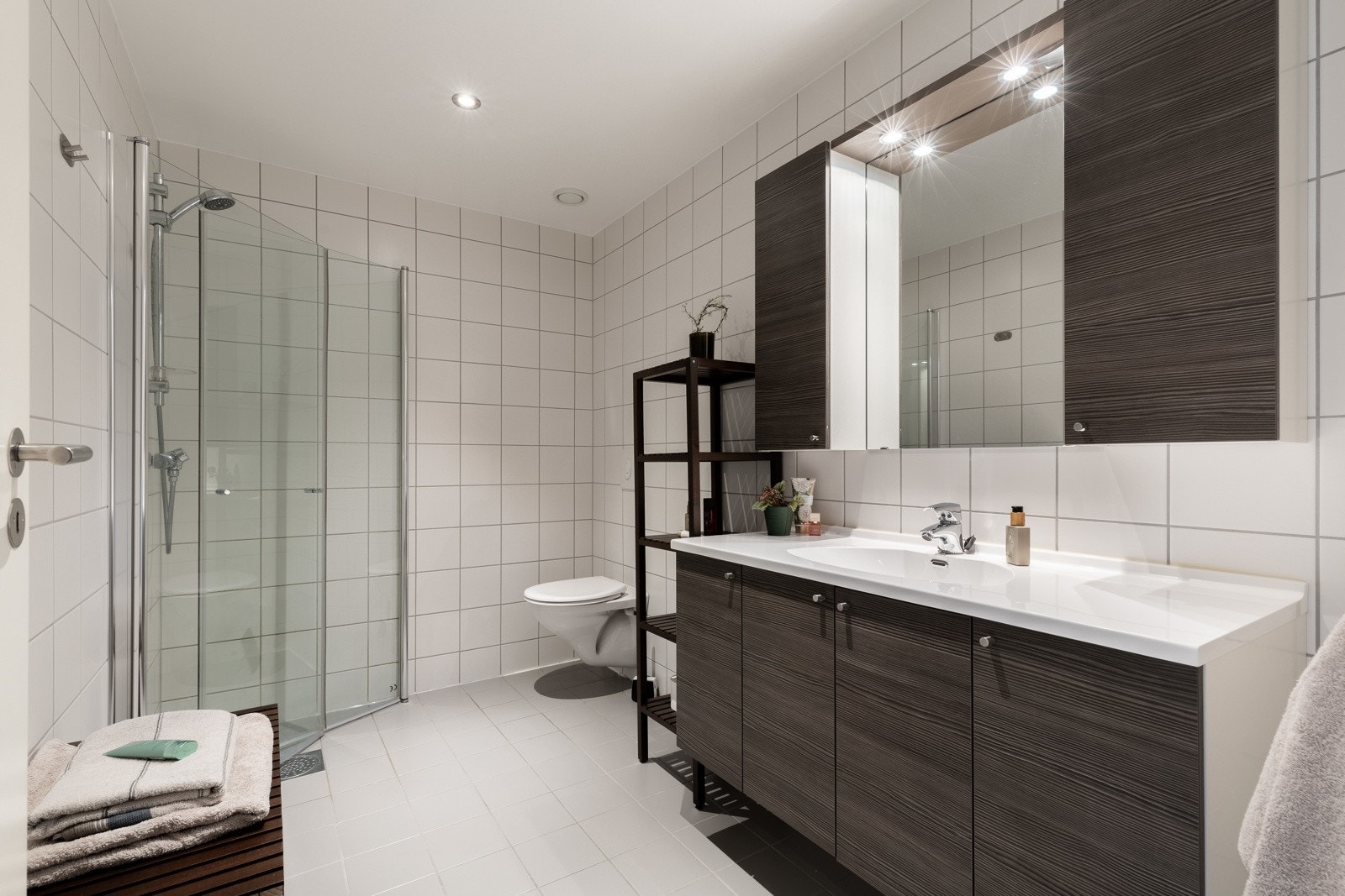 Moderne baderom med dusjhjørne, vegghengt toalett og stor servant med underskap. Lyse fliser på vegger og gulv. Galleribilde