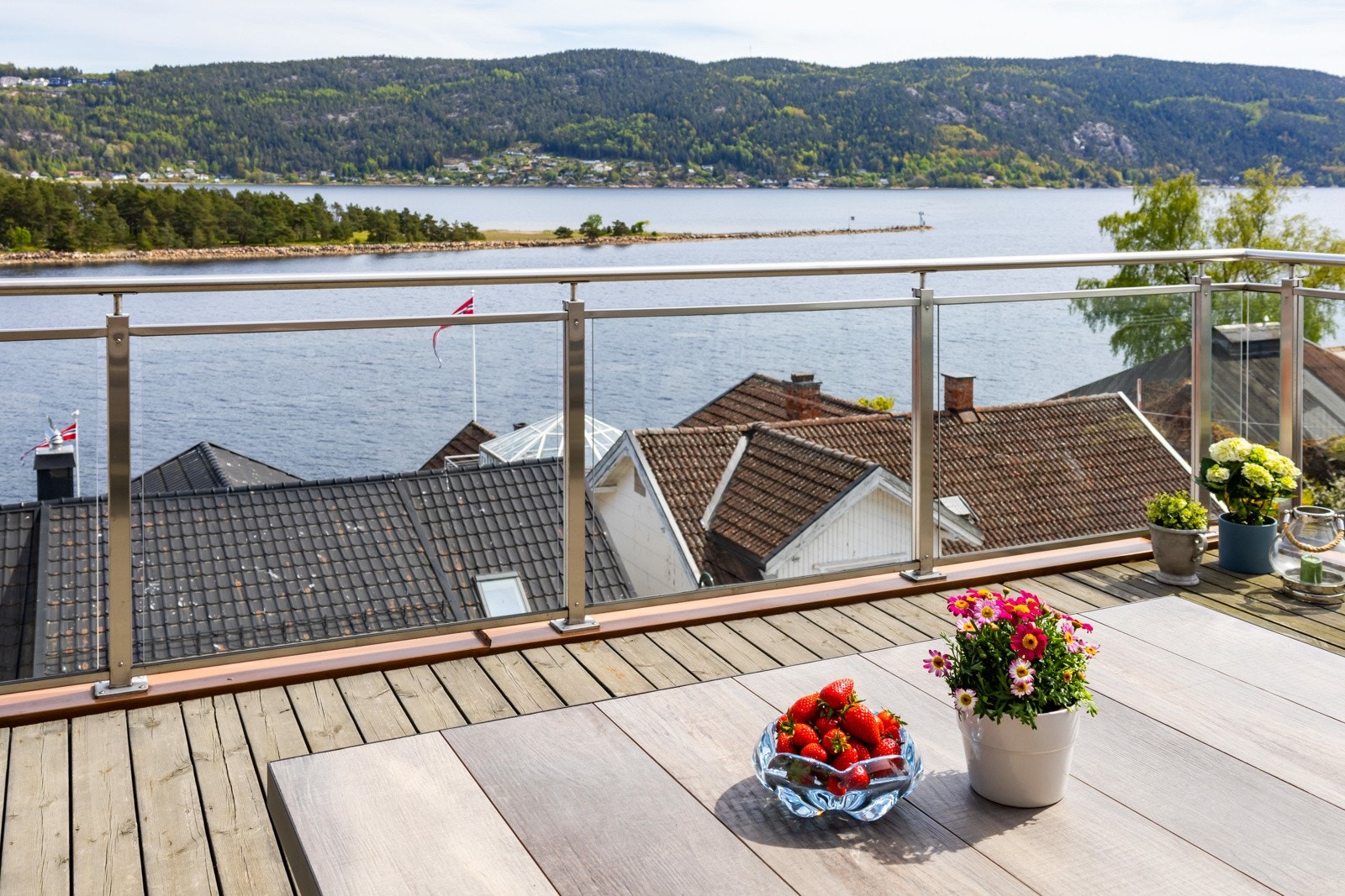 Terrasse med glassrekkverk og utsikt over vannet og skogkledde åser. Galleribilde