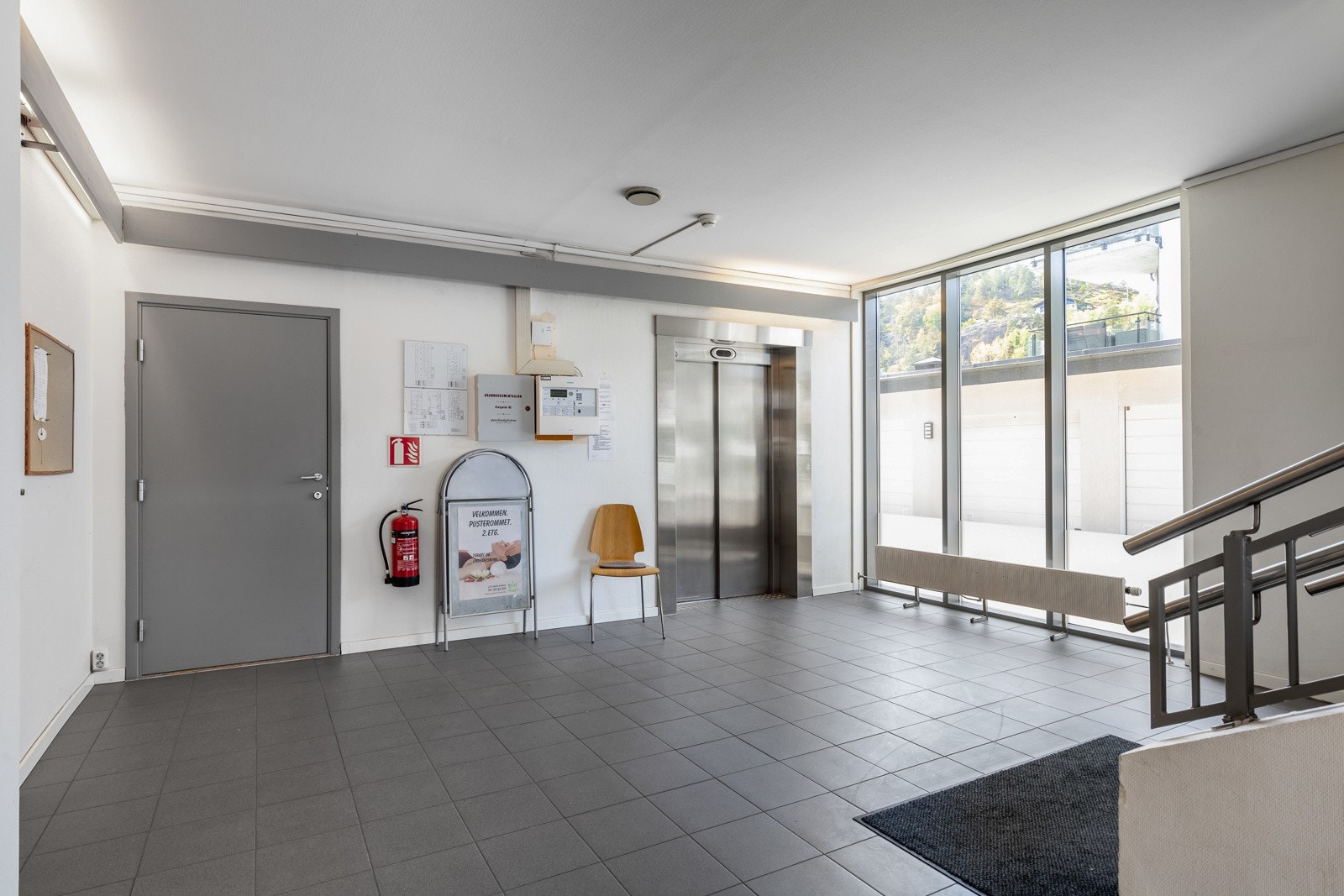 Inngangsparti med heis, fliser på gulv, store vinduer og informasjonstavle. Galleribilde