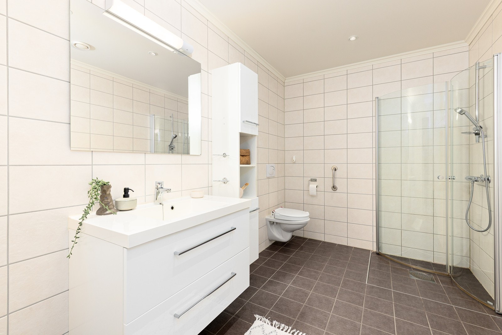 Det flislagte badet er tidløst og romslig, med varmekabler i gulvet som skaper behagelig komfort, og er innredet med nedfelt servant, høyskap, veggmontert toalett og et dusjhjørne med innfellbare glassdører. Galleribilde