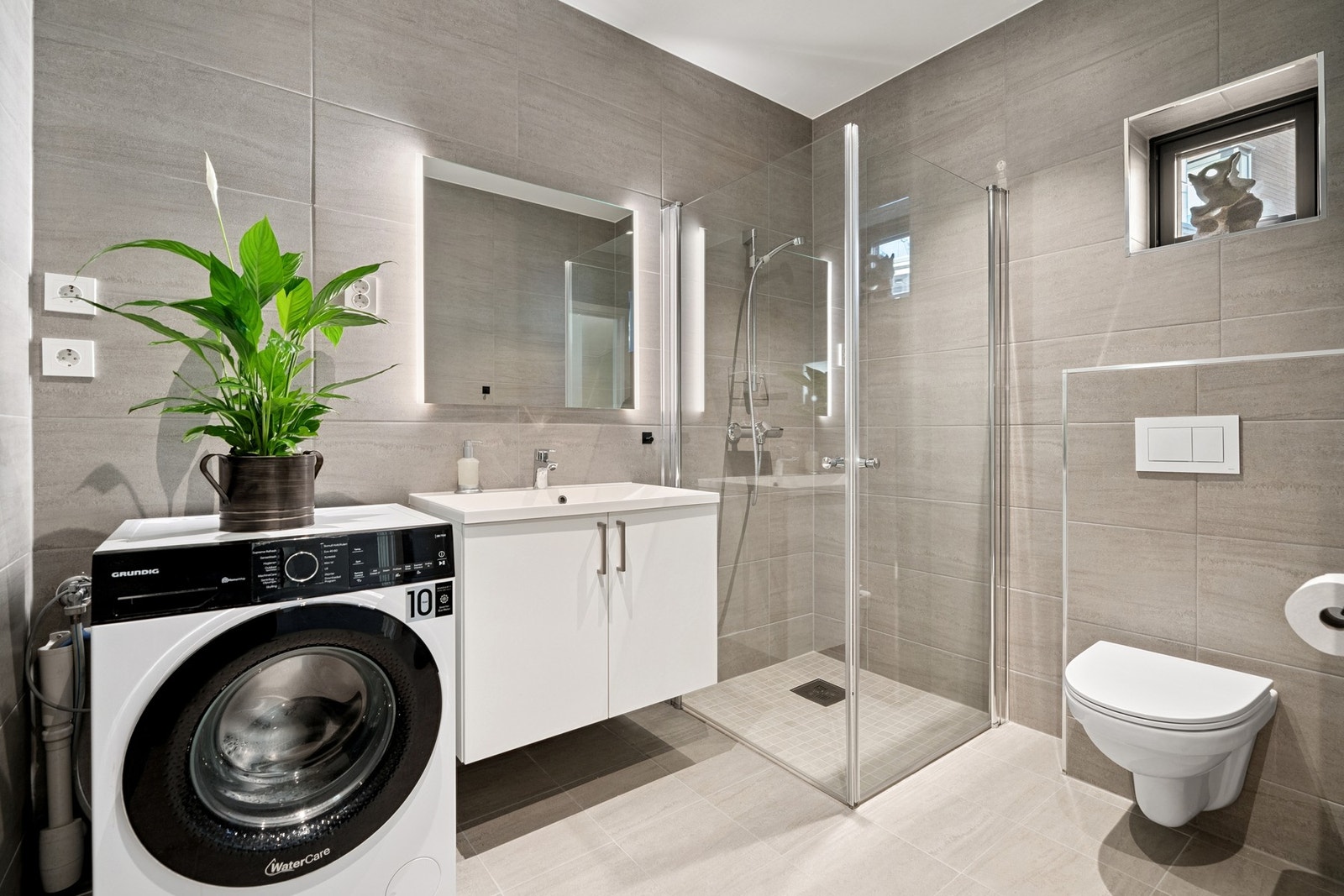 Flislagt bad med gulvvarme, vegghengt toalett, dusjhjørne og baderomsinnredning med servant. Opplegg for vaskemaskin. Galleribilde