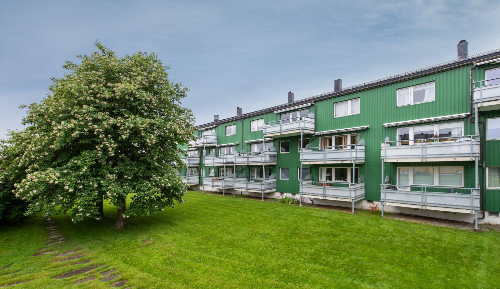 Fasade balkonger sommerstid. Rolig og fin beliggenhet i feltet med store grøntarealer mellom byggene. Parkering i gate eller på felles oppstillingsplasser, plass med elbil lader/ garasje etter ansiennitet. Galleribilde