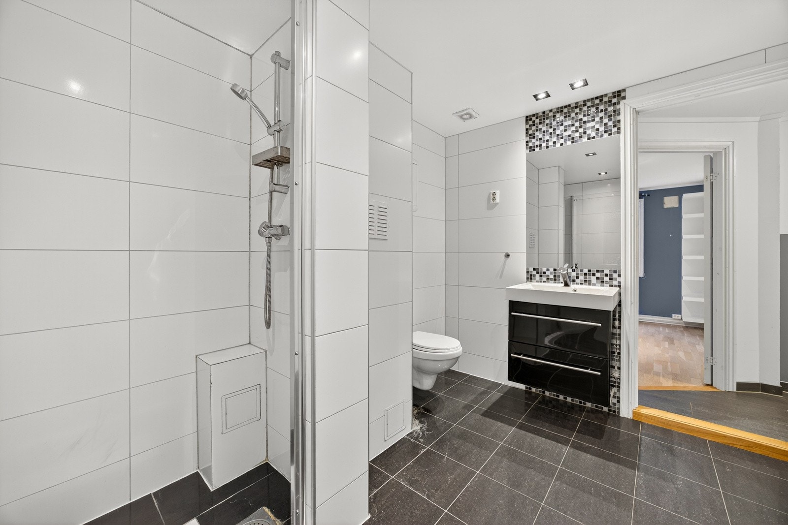 Videre har badet vegghengt toalett med innebygget sisterne og dusjhjørne med dør i herdet glass. Det er montert spottere i taket. Opplegg for vaskemaskin. Galleribilde