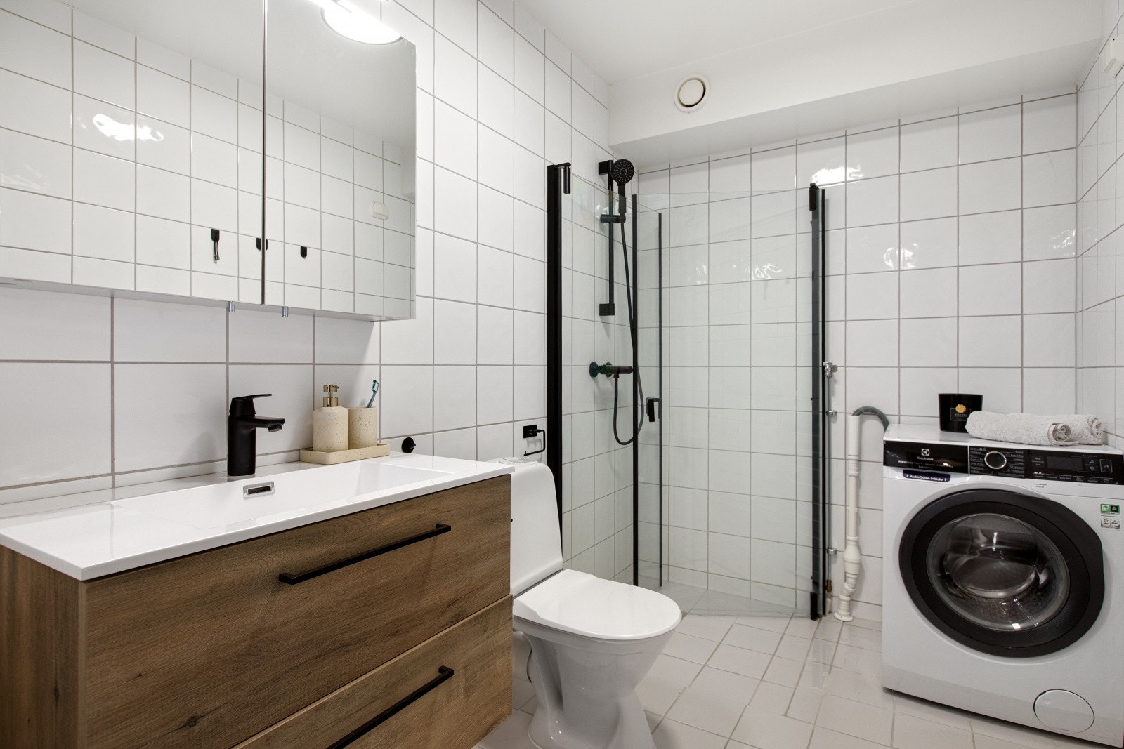 Baderomsinnredning med skuffeseksjon, servant, speilskap, toalett og dusjhjørne med innfellbare glassdører. Det er opplegg for vaskemaskin og tørketrommel på badet. Galleribilde