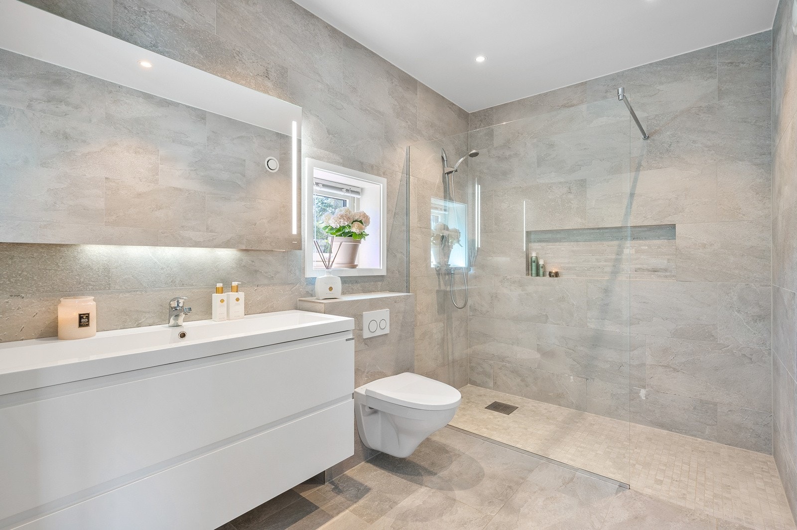 Delikat, flislagt baderom med smakfull innredning , vegghengt toalett og stor dusj med innfliset hylle Galleribilde