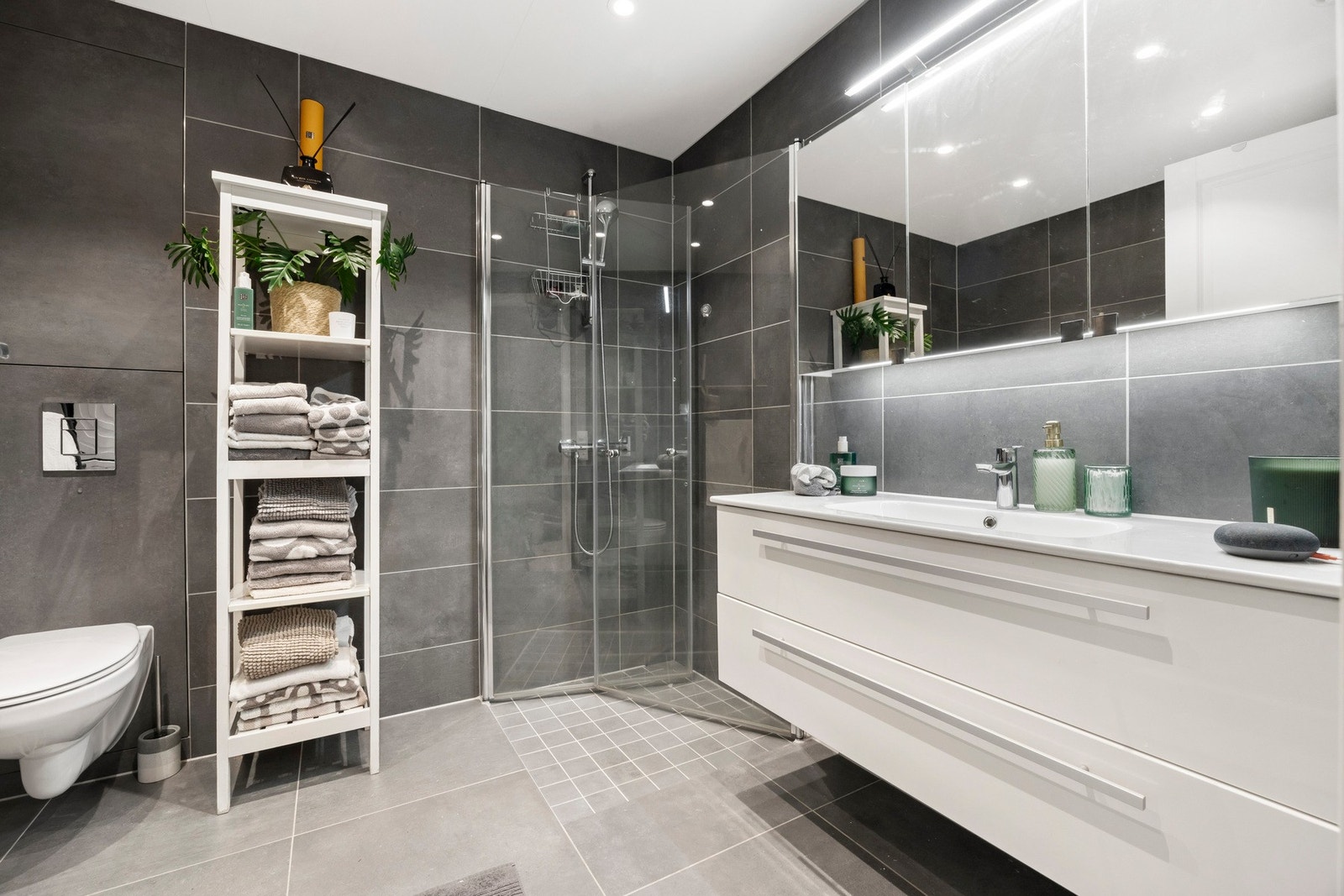 Lekkert baderom med god plass. Store fliser på gulv og vegger samt dusjhørne med glassdører, servant og servantinnredning med skuffer og speil med belysning over servanten, vegghengt toalett og downlights Galleribilde