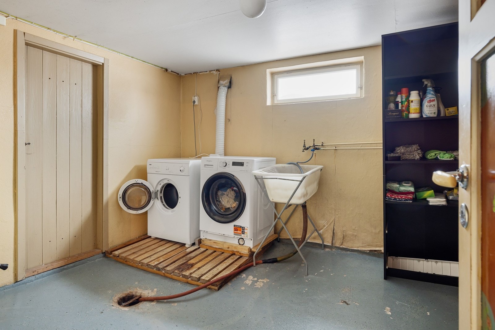 Vaskekjeller fra byggeår uten fremlagt dokumentasjon. Rommet har malt/behandlet betong på gulv, vegger av tapetsert og malt mur, samt vedovn som varmekilde. Galleribilde