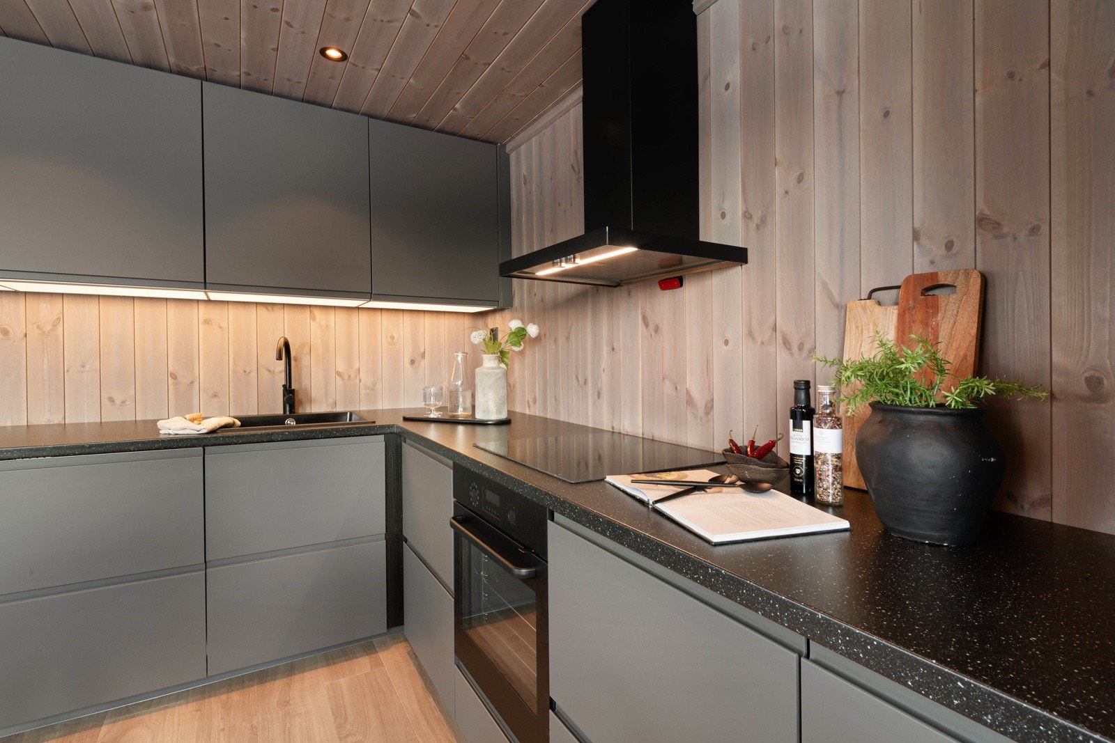 Den har takhøye skap, grå, glatte fronter og benkeplate i sort laminat med nedfelt vaskekum i sort kompositt. Galleribilde