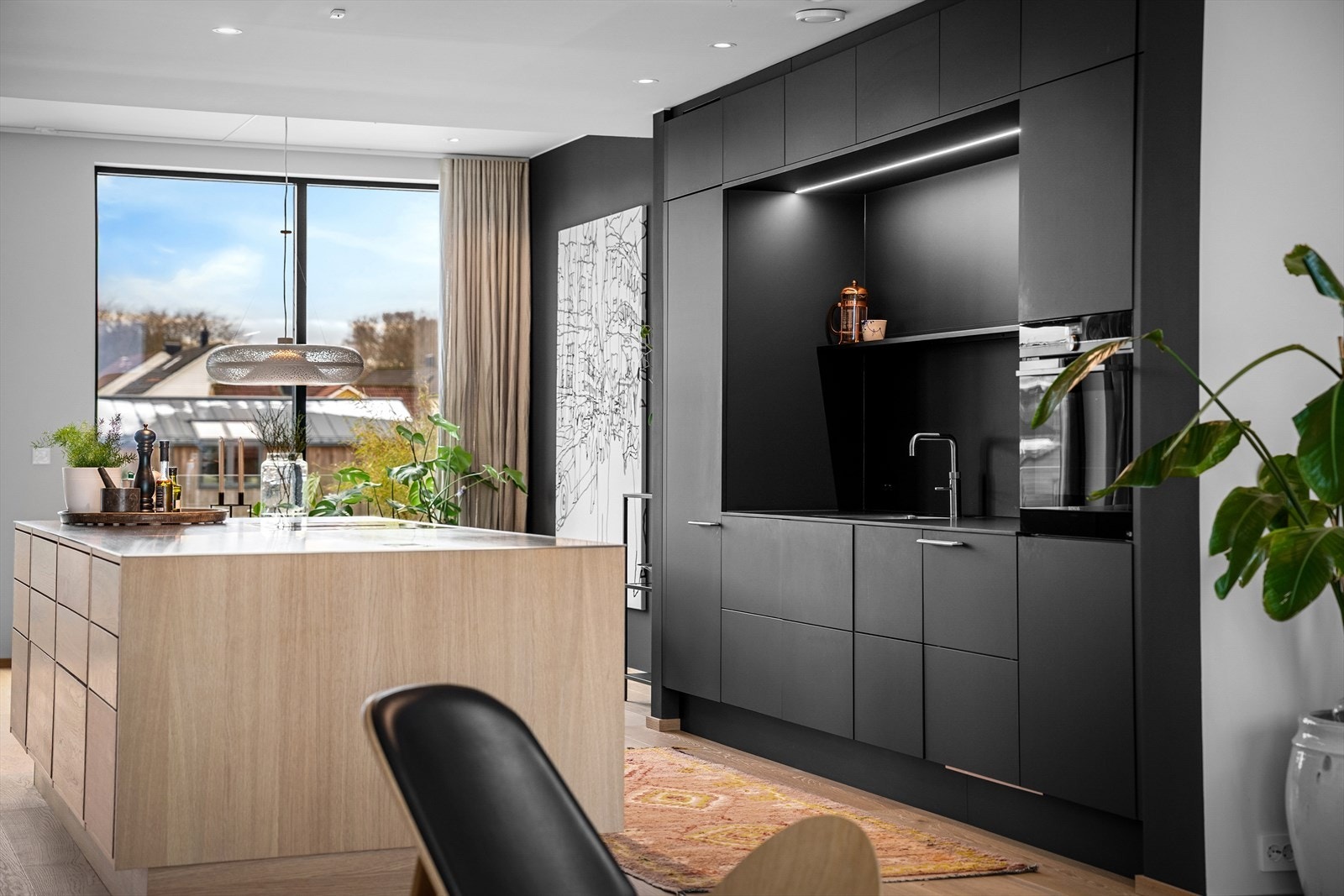 Det harde mot det myke. Sorte fronter og slank, stål benkeplate på øy i lun kontrast til eikeskuffer. Led belysning over planlimt vask med Qooker, og sort benkeplate som glir sømløst i ett med resten. Gjennomgående naturlig lys fra vinduer. Galleribilde