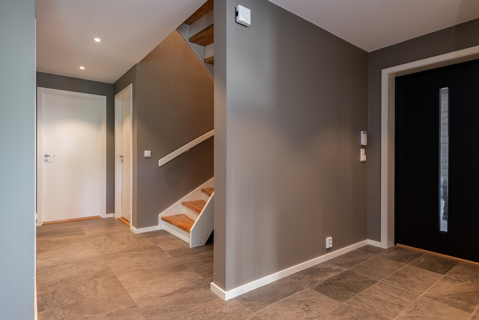 Flislagt gulv i entréen gir et robust og praktisk inngangsparti. Trappen har en moderne utførelse med tretrinn og mangler rekkverk på nedre del, noe som bør vurderes for sikkerhet. Galleribilde