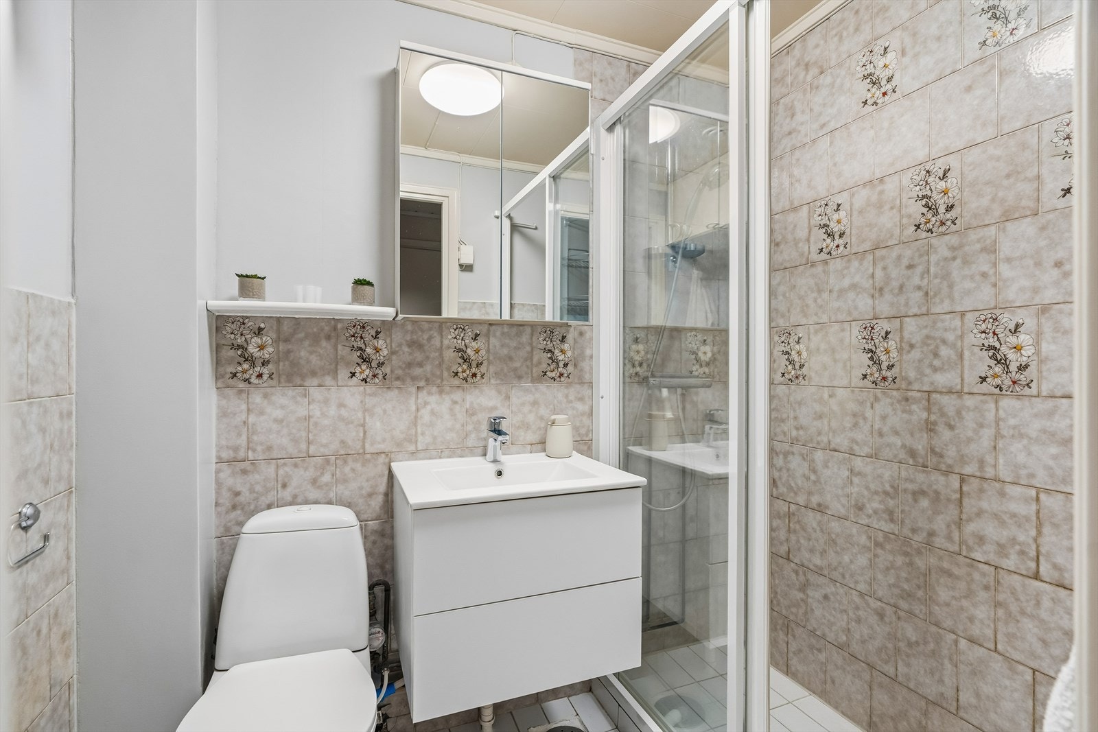 Det er inngang til et flislagt baderom. Rommet har dusjhjørne, servant med underskuffer samt gulvstående toalett. Galleribilde