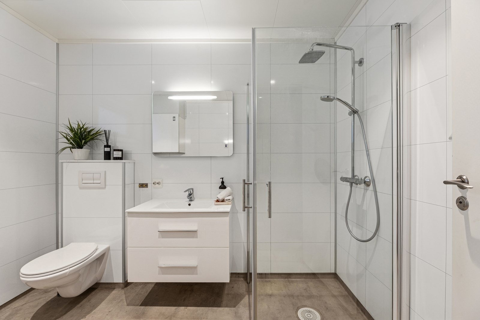 Rommet er innredet med servantseksjon, veggmontert toalett og et stilrent dusjhjørne med glassvegger. Det er også lagt til rette for vaskemaskin, noe som gjør rommet både praktisk og funksjonelt. Galleribilde