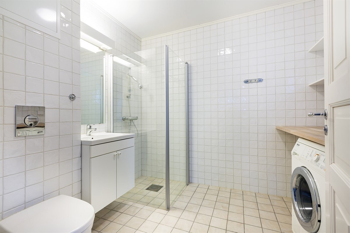 Romslig og delikat bad med lyse fliser og moderne detaljer. Vegghengt servantskap og toalett gir et stilrent uttrykk, mens hjørnedusjen med glassdører sørger for god plassutnyttelse og et luftig preg. Galleribilde