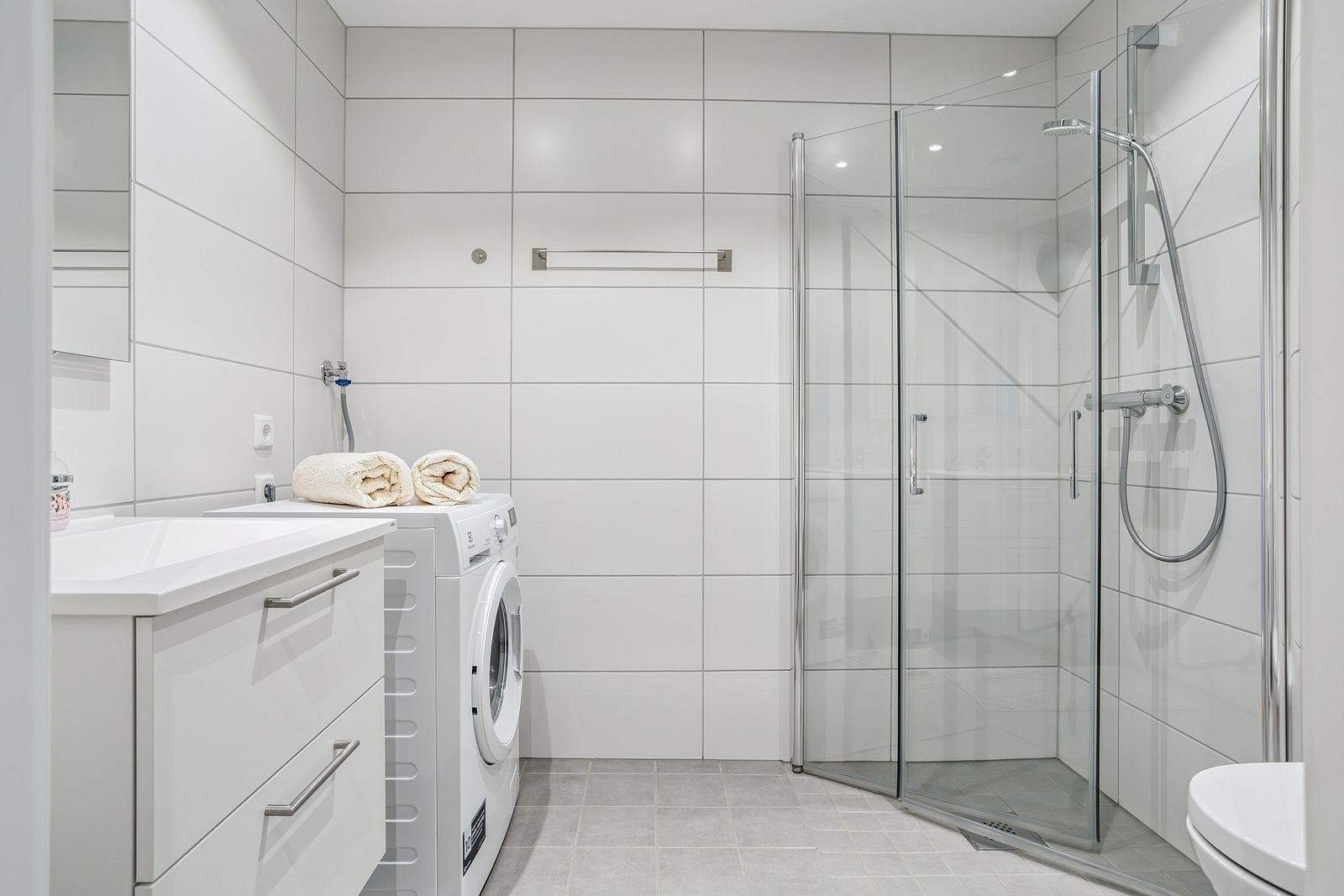 Badet er lyst og lekkert og inneholder vegghengt toalett, dusj, servant og kombimaskin Galleribilde