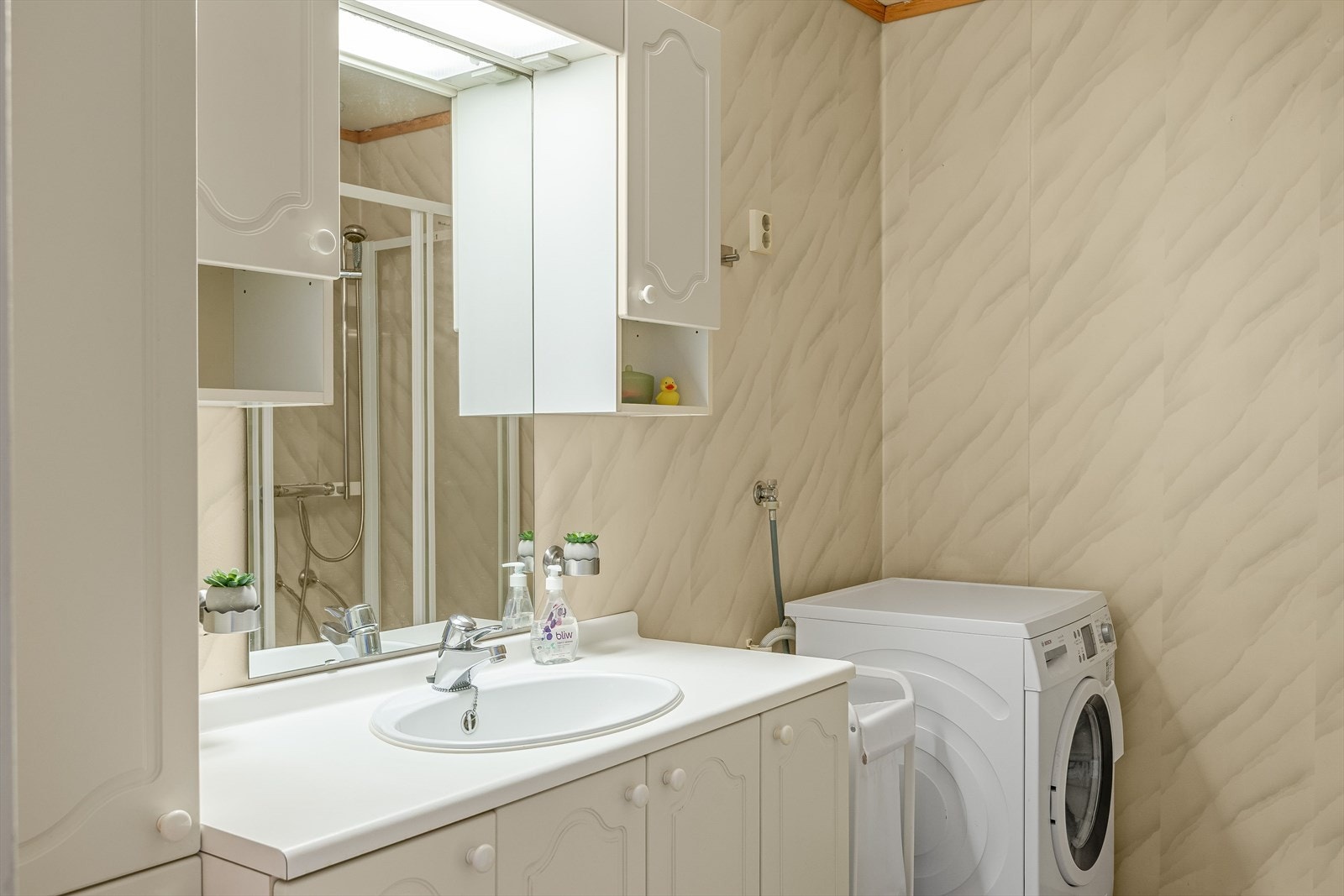 Lys baderomsinnredning med profilerte fronter og laminert benkeplate med nedfelt servant, speil med belysning og vegghengte skap over. I hjørnet er det plass og opplegg til vaskemaskin, samt tørketrommel over om ønskelig. Galleribilde