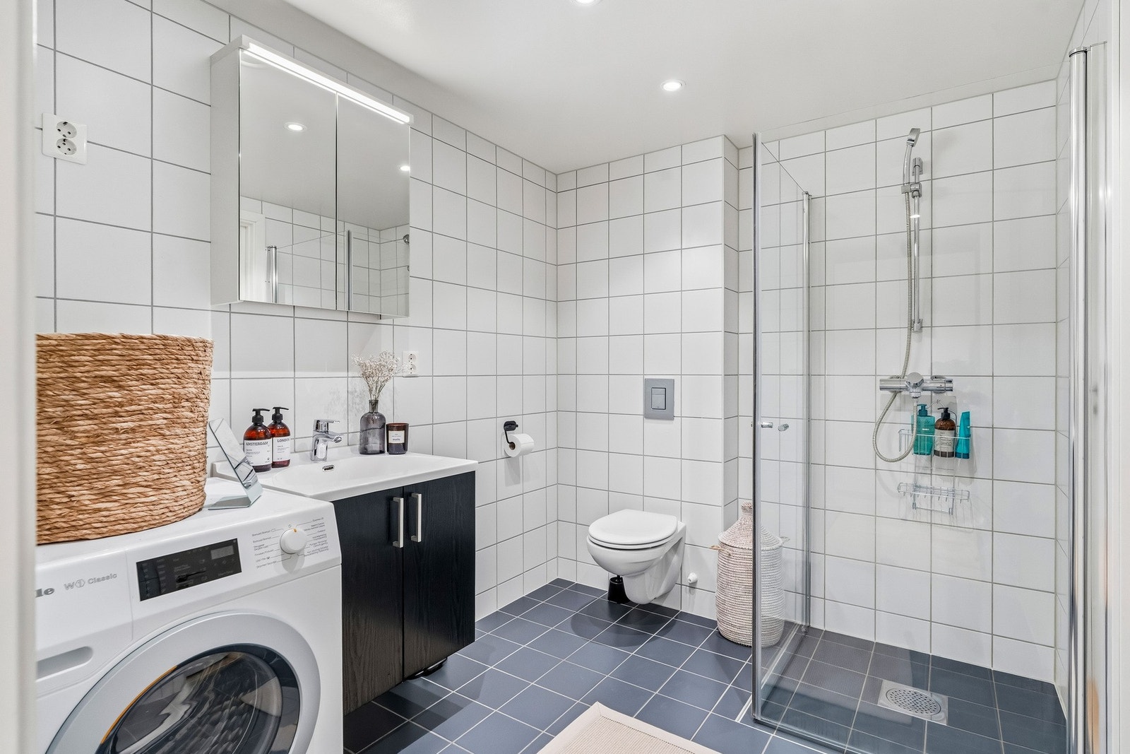 Flislagt bad med vannbåren gulvvarme og spotter i himlingen. Baderomsinnredningen består av benkeskap med glatte fronter og veggskap med speilfronter. Badet er utstyrt med dusj med innfellbare dører i herdet glass, servant samt veggfestet klosett med inne Galleribilde