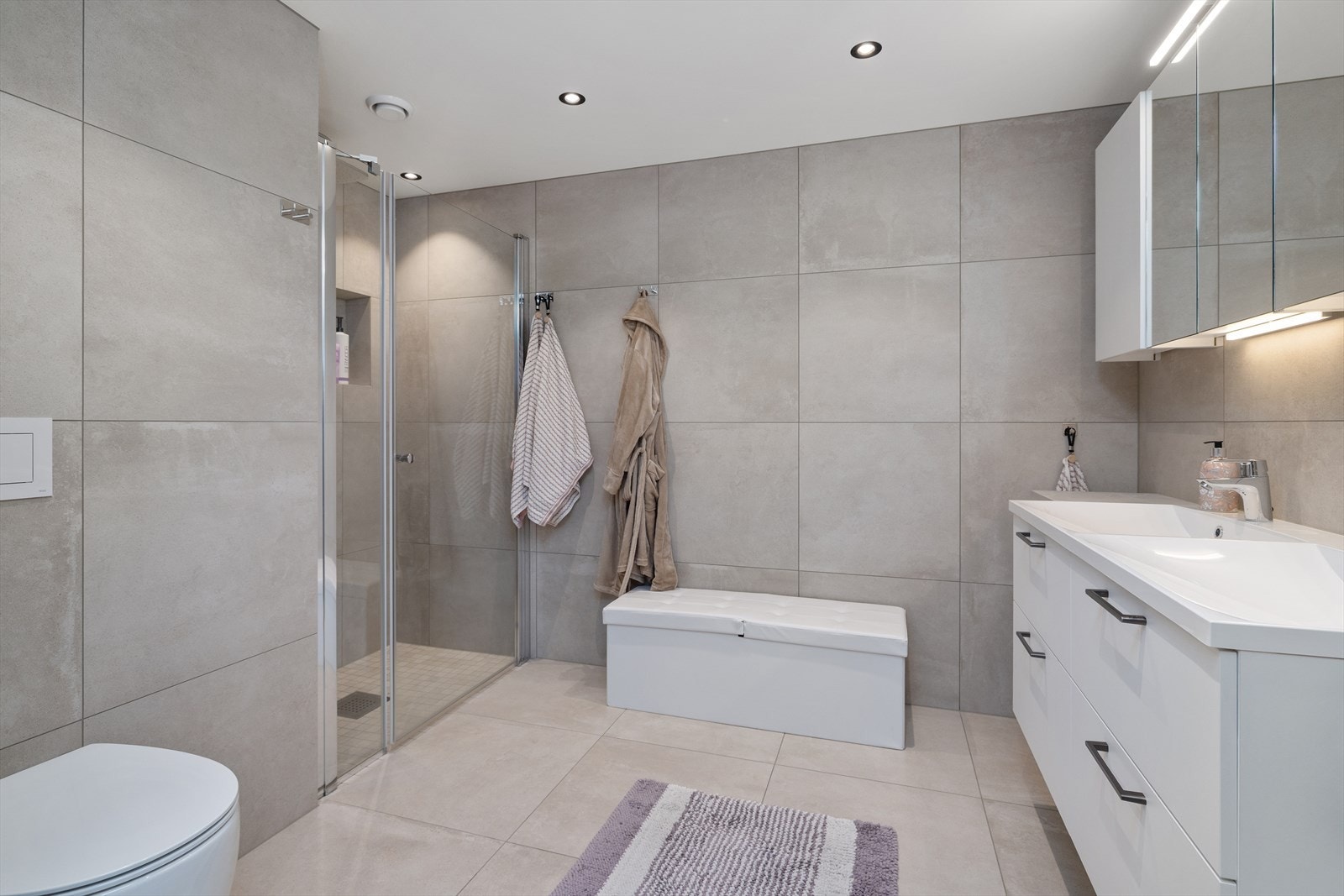 Badet har veggmontert toalett og dusjvegger/hjørne. Det er balansert ventilasjon. Spotter i taket, fliser på gulv og vegger. Galleribilde