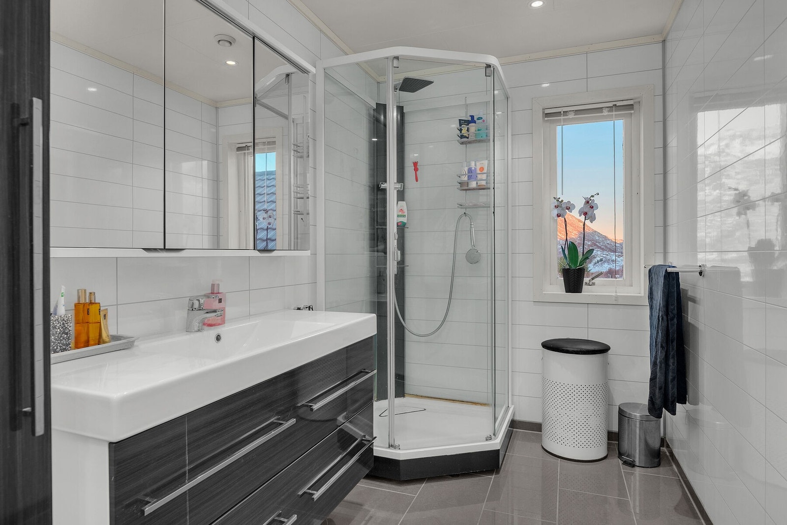 Badet er innredet med servant på skuffeseksjon, høyskap, speilskap, dusjkabinett og veggmontert toalett. Galleribilde