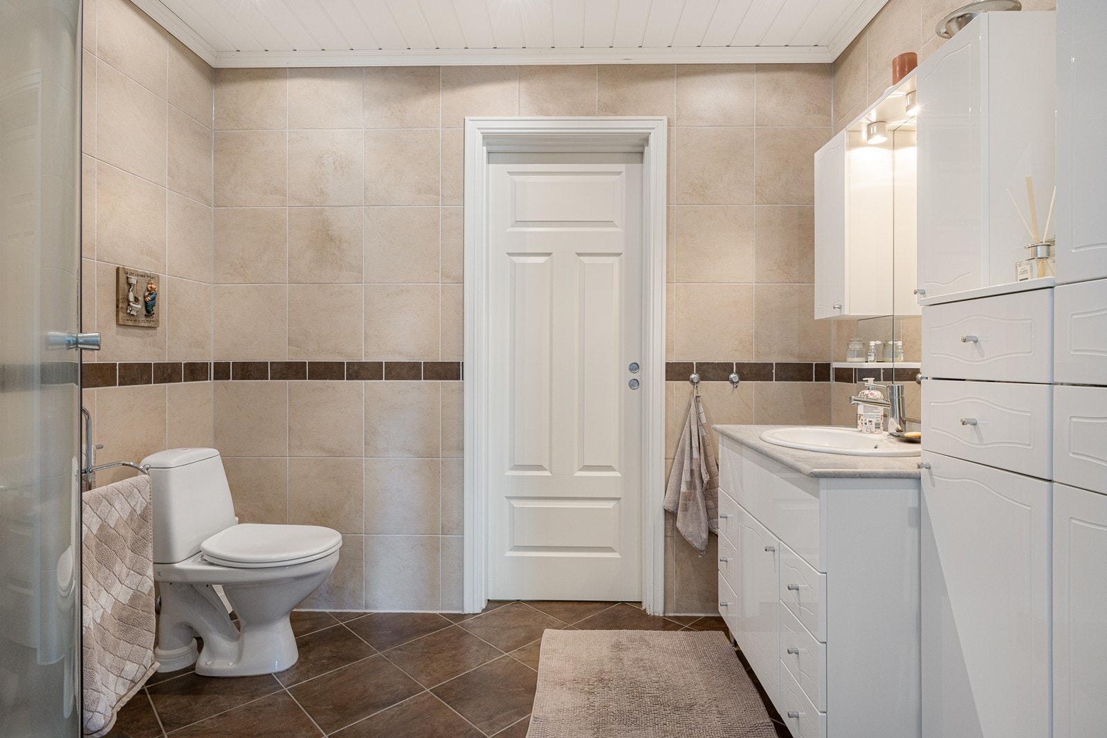 Baderomsinnredningen har hvite fronter, lysegrå benk og nedfelt servant, og over er det et stort speil med god belysning. Videre har badet et toalett og et dusjkabinett. Galleribilde