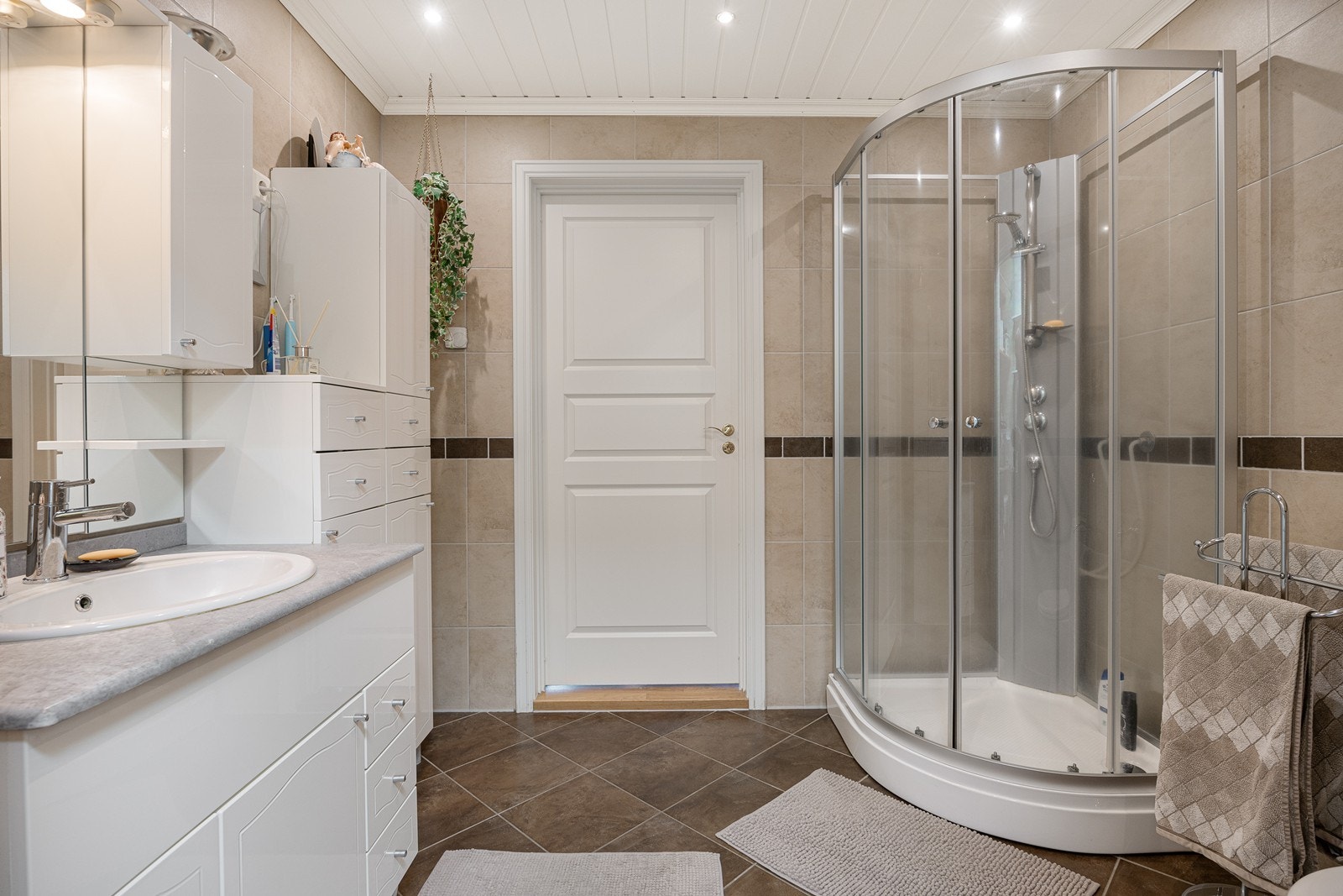 Badet fremstår som velholdt og pent, med beige fliser på veggene og brune fliser på gulv med gulvvarme. Galleribilde