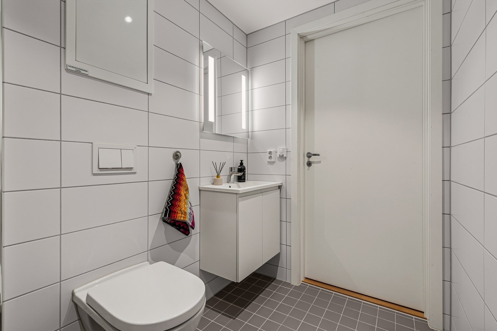 Badet er utstyrt med veggmontert toalett, dusjhjørne med to svingbare glassdører, høyskap, samt baderomsinnredning med servant, blandebatteri og speil. Galleribilde