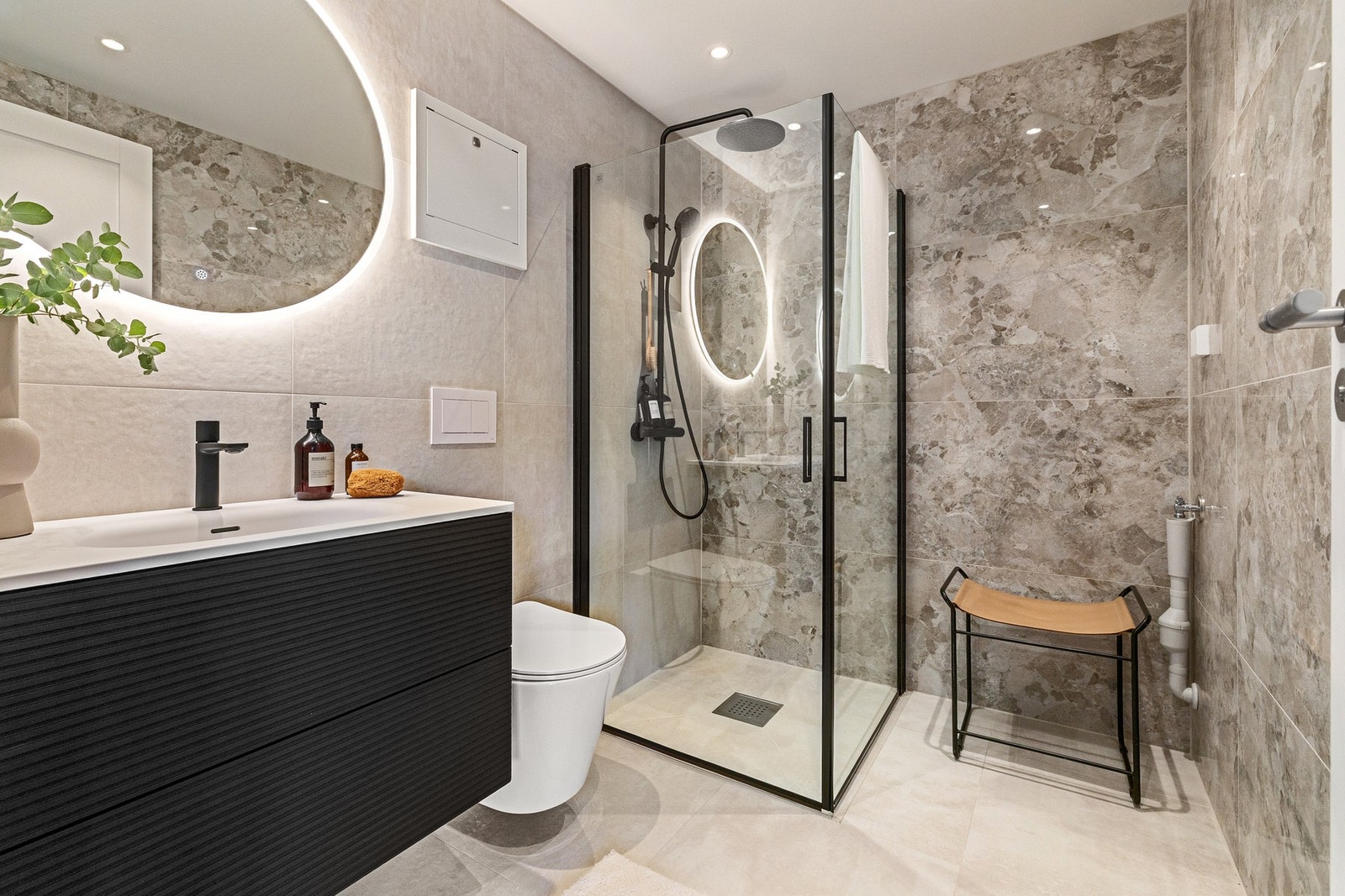 Eksklusivt bad med store fliser i elegante naturtoner. Servantskap i mørk utførelse med ovalt LED-speil, integrert lys og gulvvarme. Galleribilde