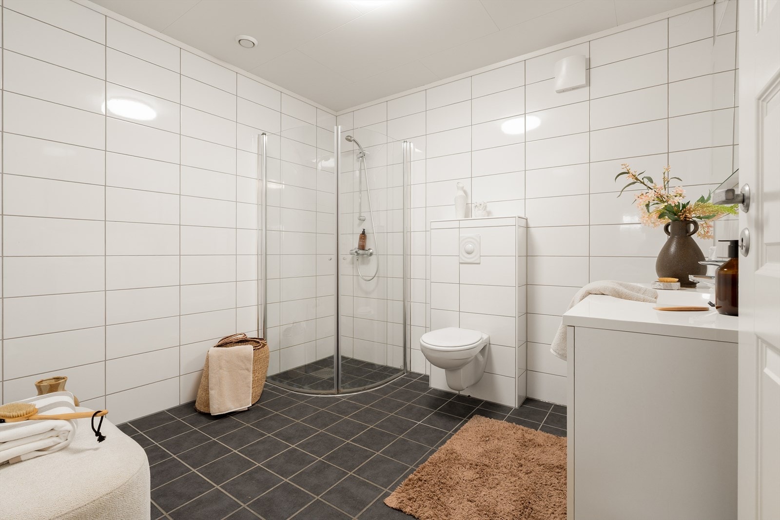 Rommet er innredet med servant i møbel, speil, vegghengt toalett og dusj med glassdører - et flott og stilrent uttrykk. Galleribilde