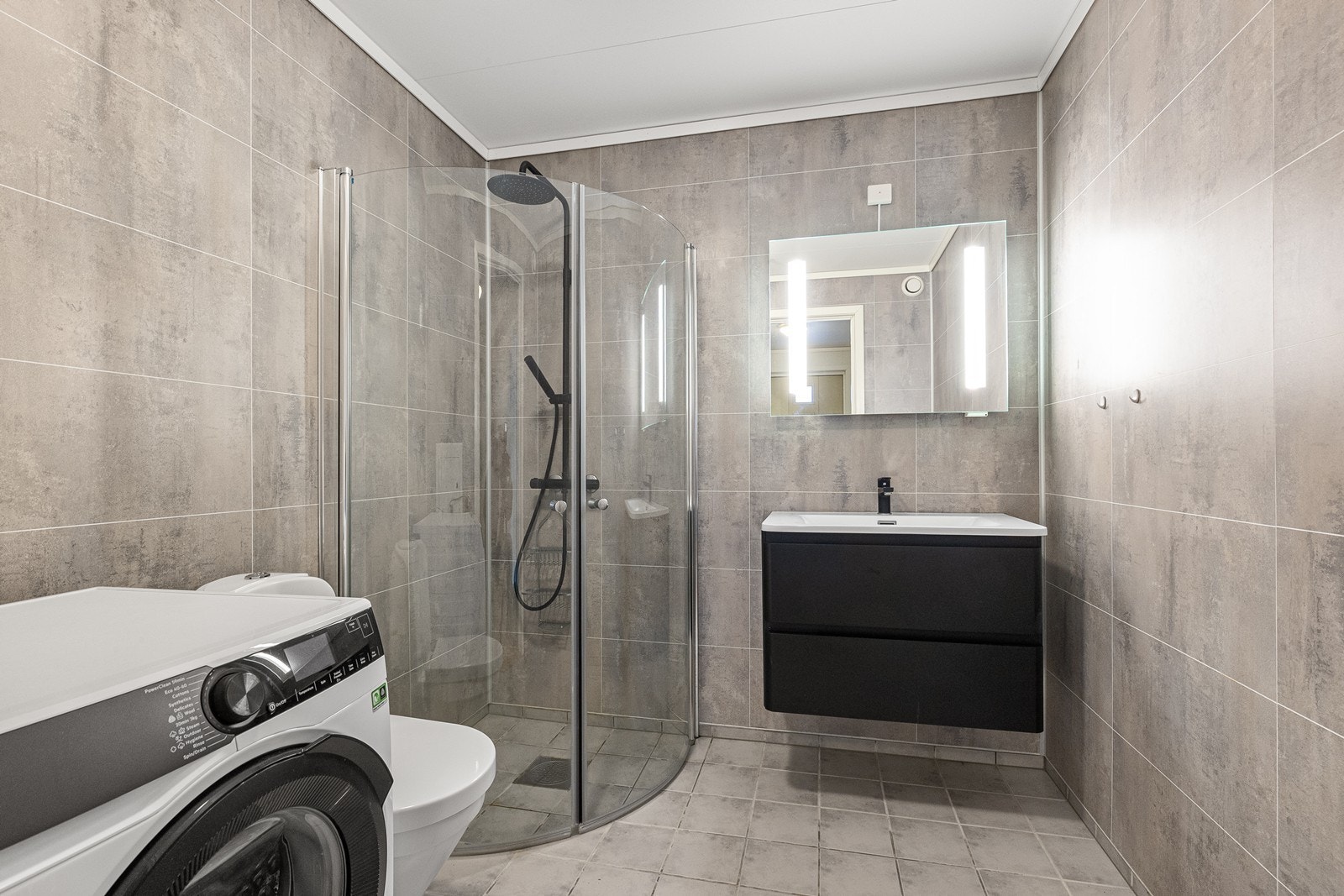 Pent bad med dusjhjørne, vegghengt toalett og opplegg for vaskemaskin. Galleribilde