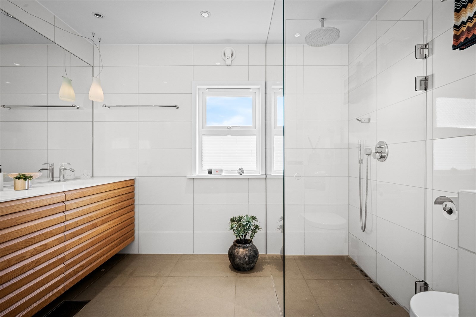 Delikat baderom med seksjon i lunt tre i kontrast til fliser. Vannfallsdusj med garnityr rett ut fra tak og vegg. Vindu for naturlig lys. Designbelysning over seksjon. Galleribilde