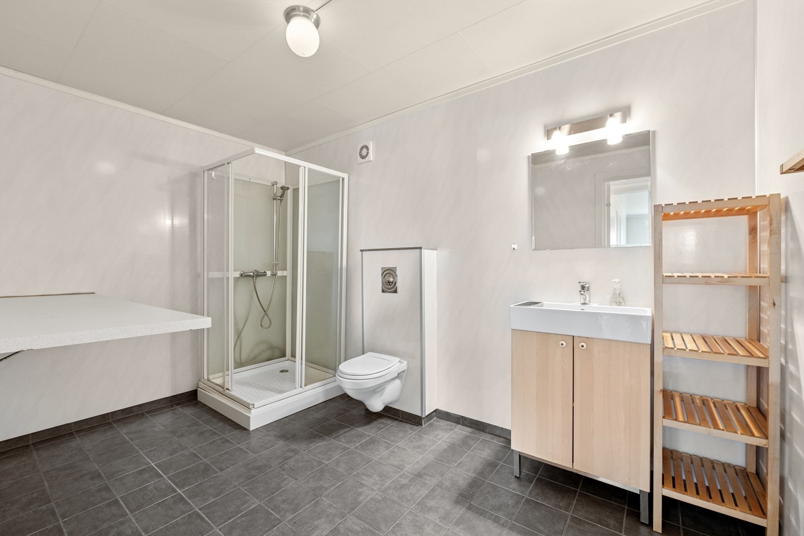 Stort baderom med flislagte gulv på betong, sluk av plast og vannbåren varme i gulvet. Vegger med baderomspanel og takessplater i himlingen. Rommet inneholder innredning med servant, vegghengt WC, dusjkabinett og opplegg for vaskemaskin og tørketrommel. Galleribilde