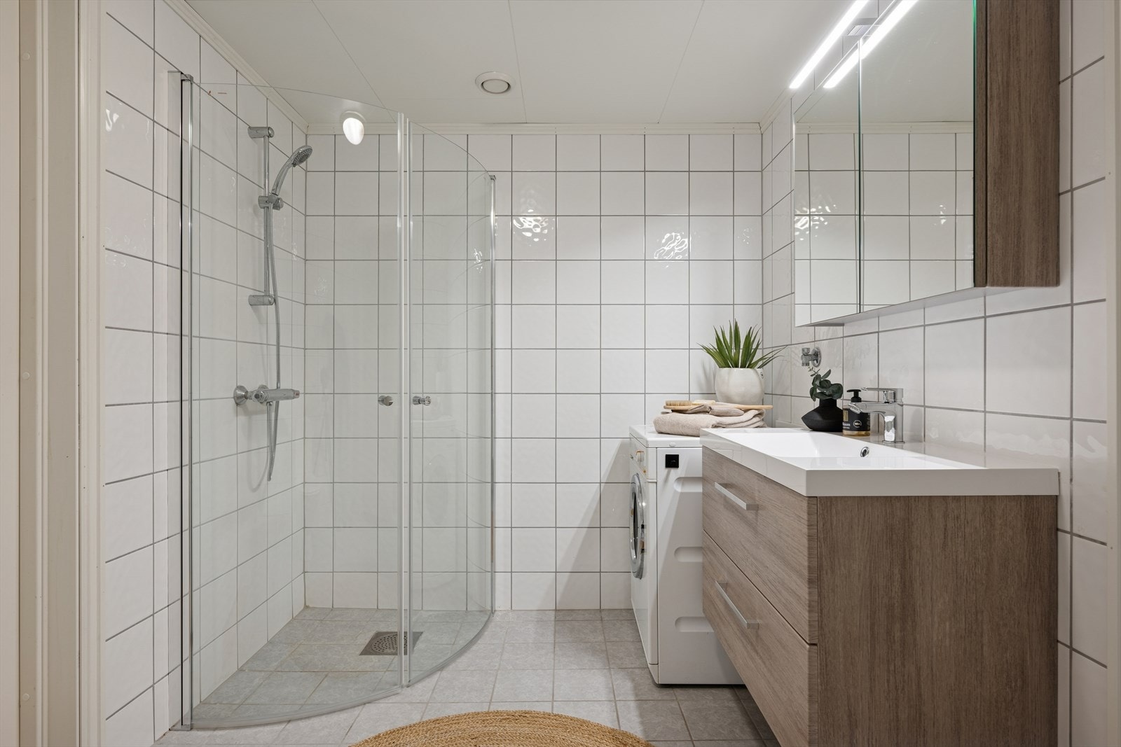 Pent flislagt bad med dusjhjørne, gulvstående toalett og opplegg for vaskemaskin. Galleribilde