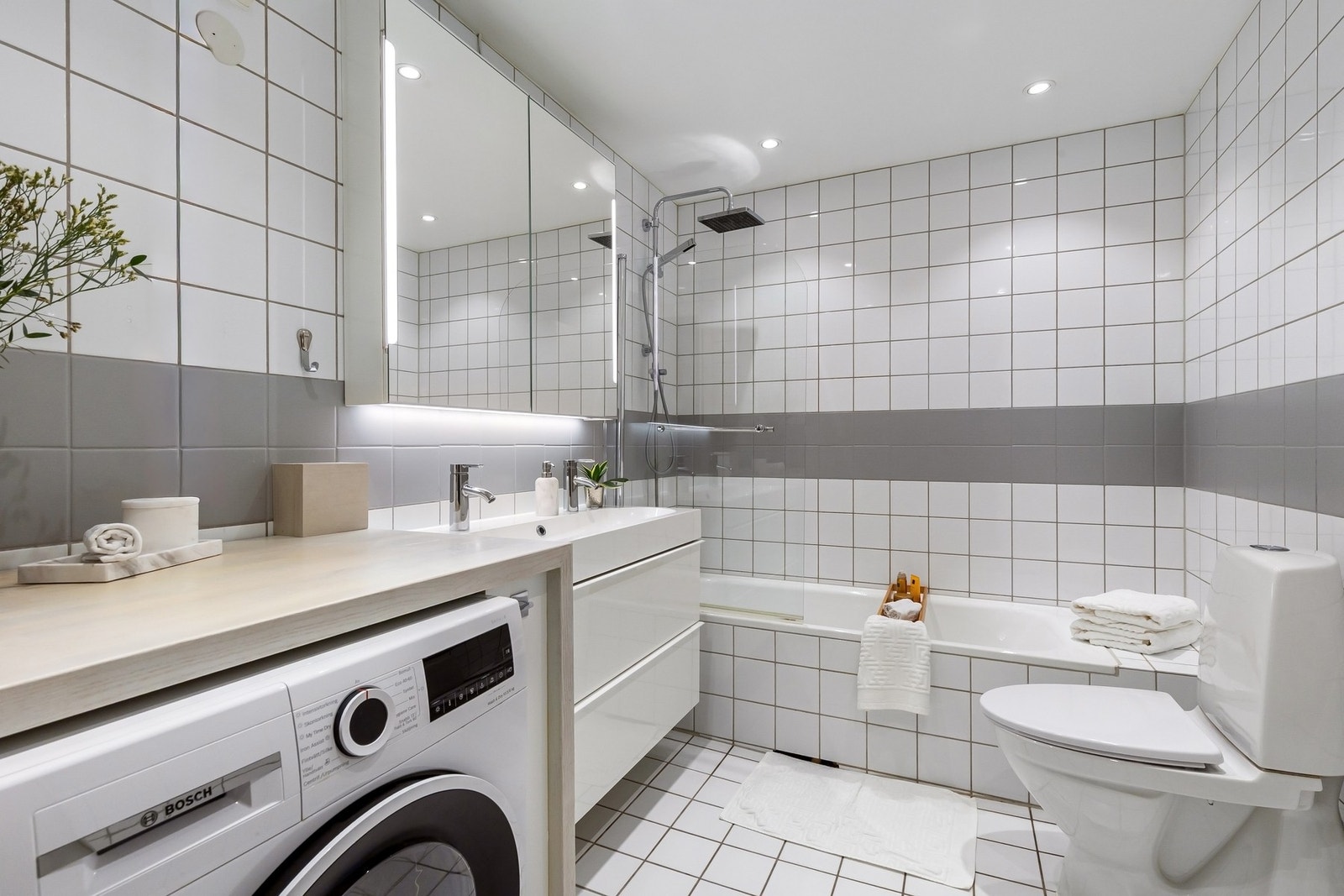 Tidløst og stort flislagt baderom med badekar og opplegg for vaskemaskin. WC og servant med underskap og vegghengt speilskap med sidebelysning Galleribilde