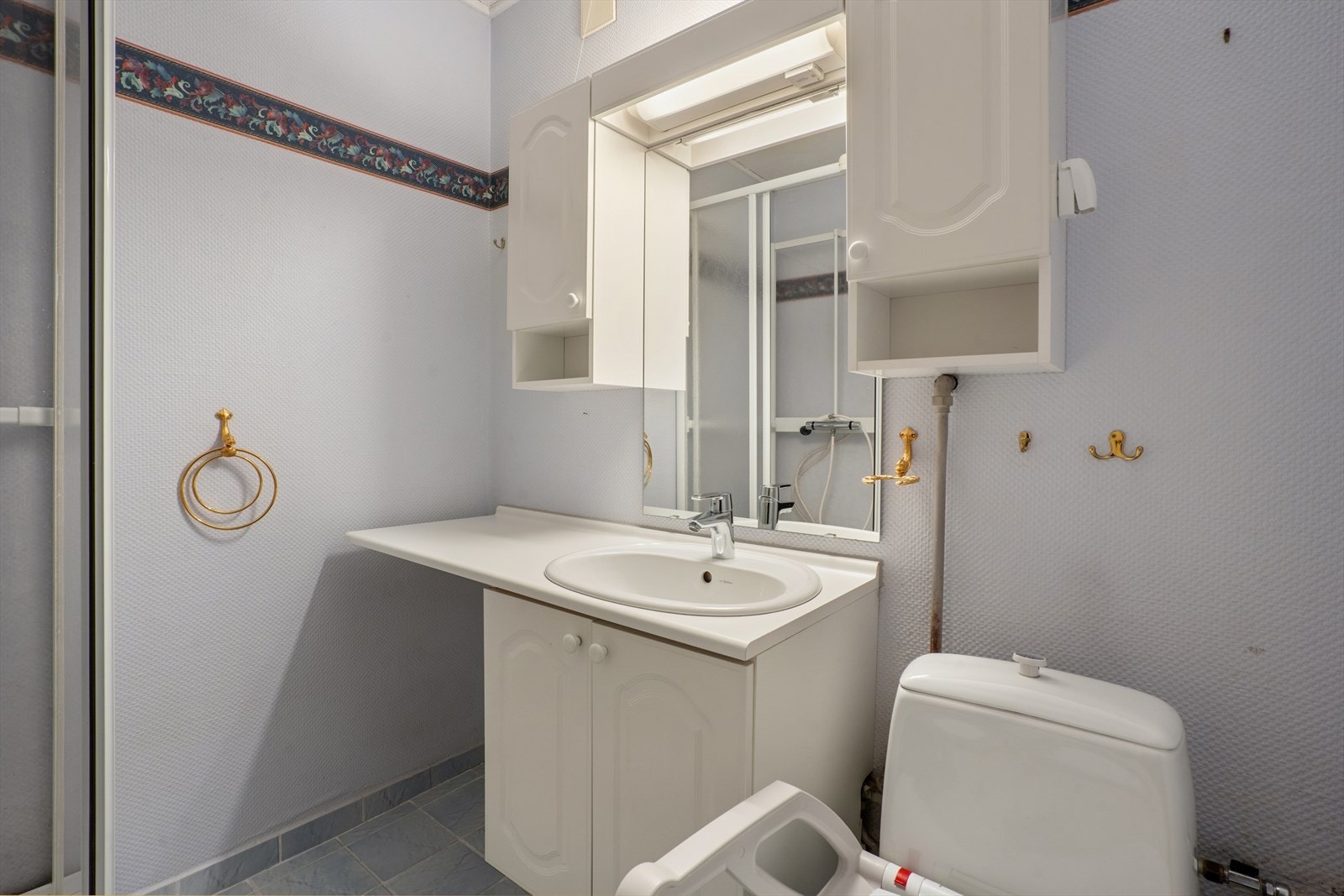 Rommet er innredet med et eldre dusjkabinett, servant med ettgrepskran og underskap, samt speil med sideskap og belysning. Galleribilde