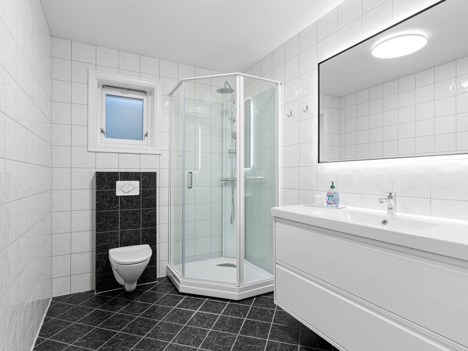 Tidløst, flislagt baderom er utstyrt med veggmontert toalett, nytt dusjkabinett og ny servantseksjon med speil. Galleribilde