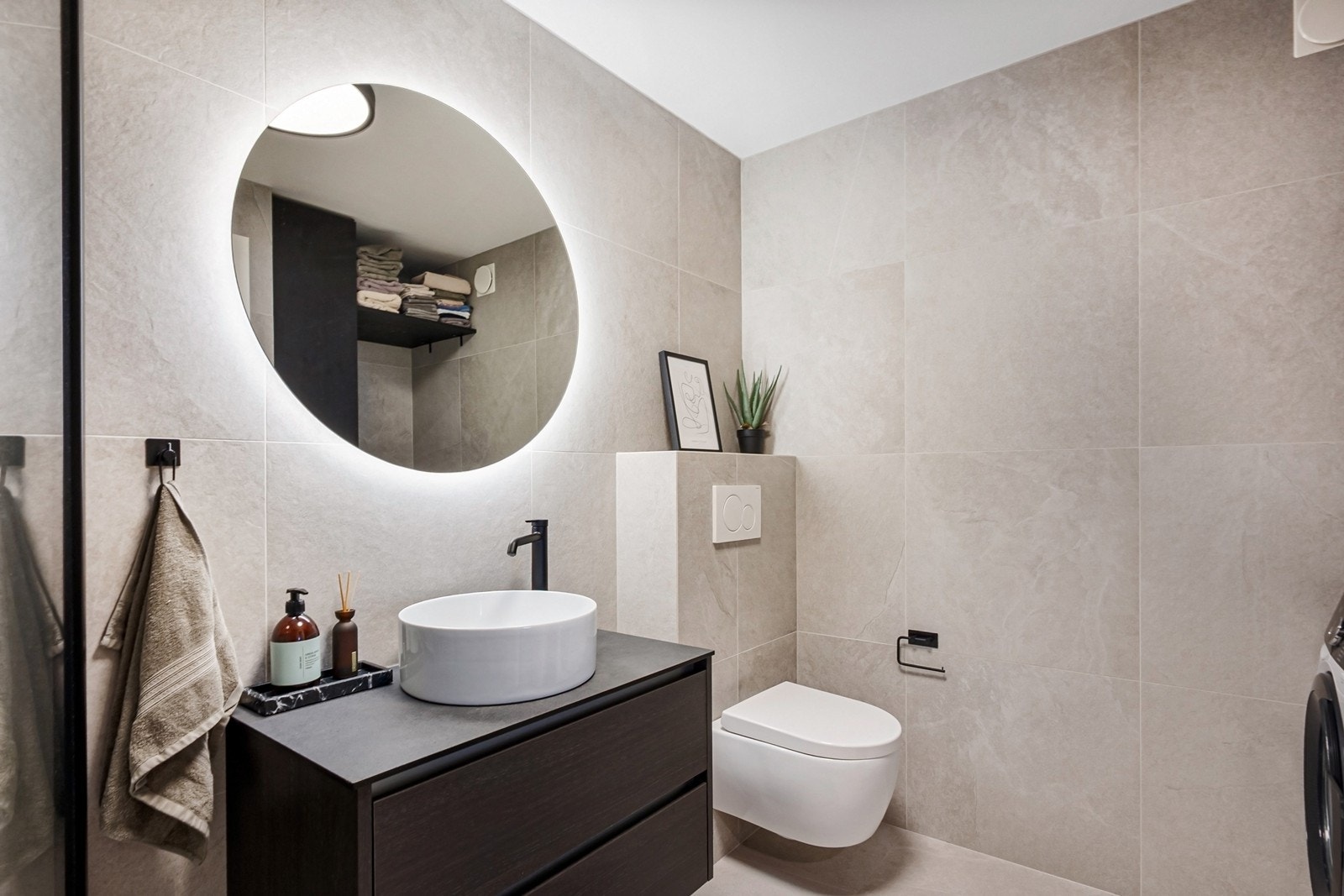 Badet har servant med skuffeseksjon og bollevask, speil og vegghengt toalett. Videre er det opplegg for vaskemaskin og tørketrommel, samt dusjhjørne med glassvegger og tak- og hånddusj i sort armatur. Galleribilde